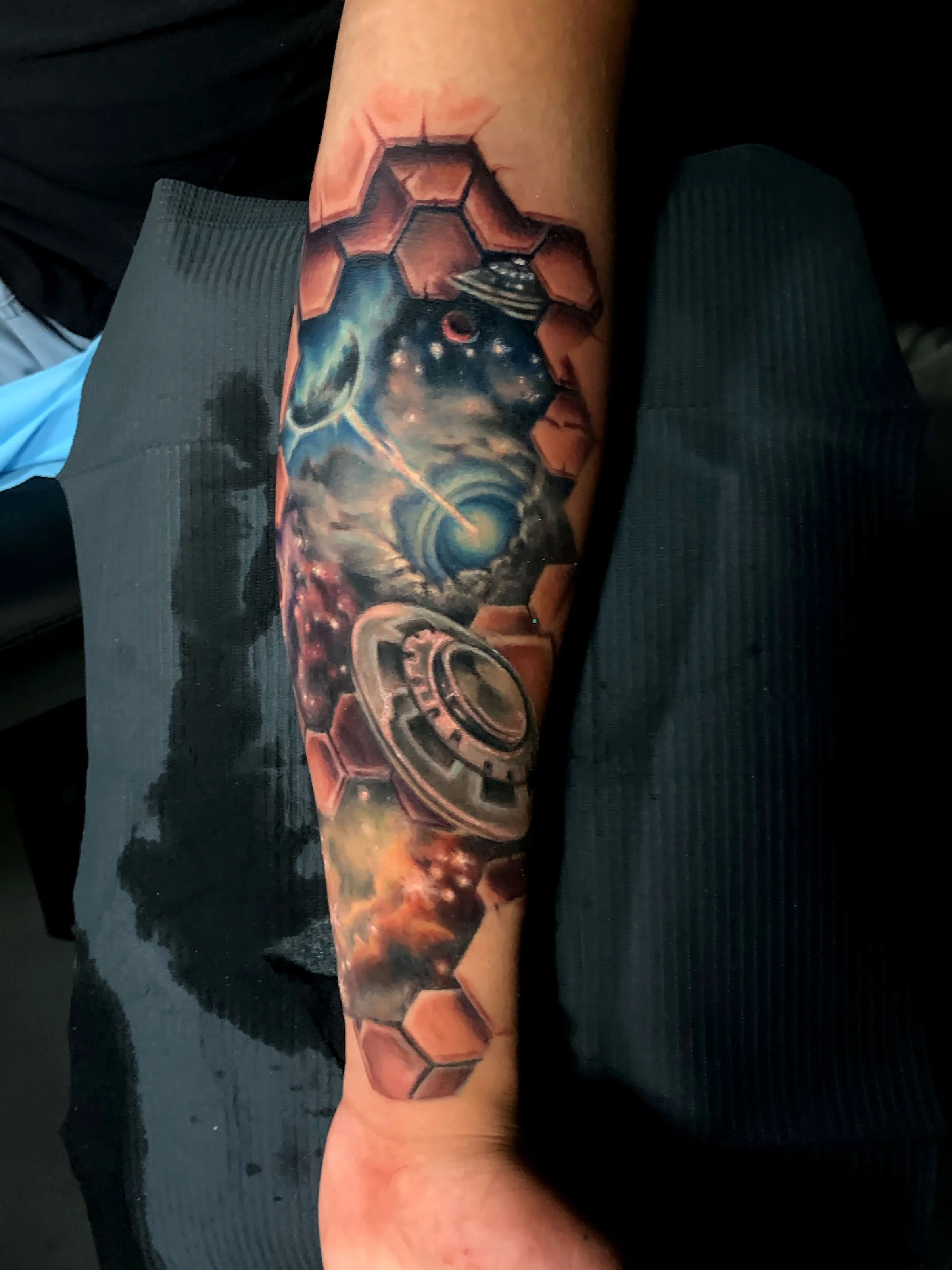 Tattoo of a galaxy, planets, and honeycomb geometric shapes on a person's arm.