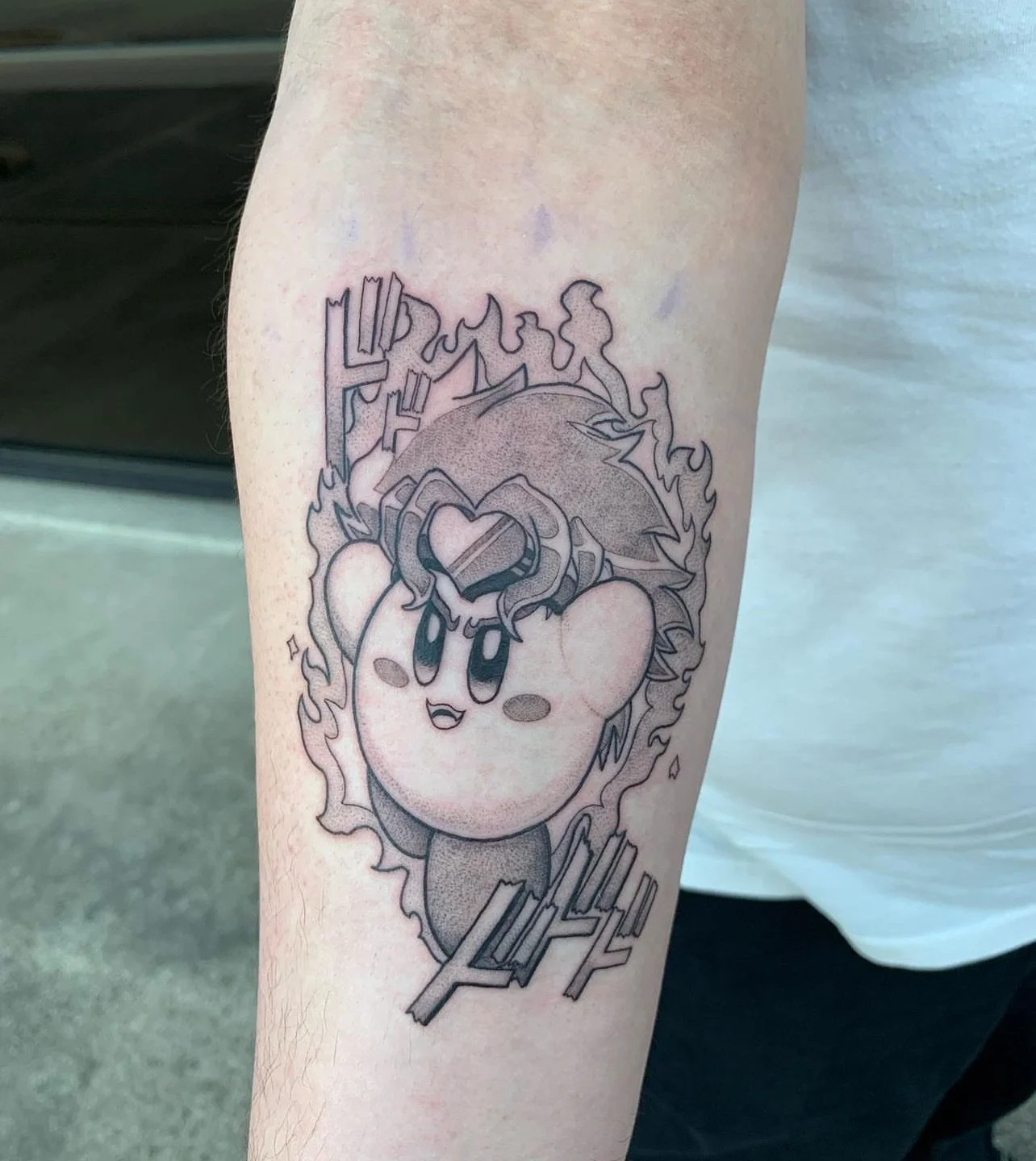 Tattoo of Kirby, a pink cartoon character, with a happy expression, surrounded by flames, with a box of ketchup on its head and Japanese-style characters below.
