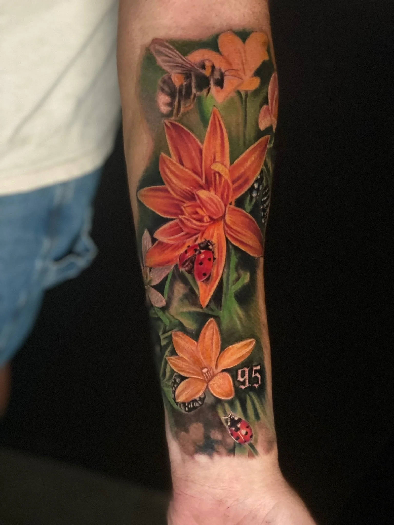 A colorful tattoo on a person's arm featuring orange lilies, a bee, a ladybug, a butterfly, a number '95', and yellow flowers against a green background.
