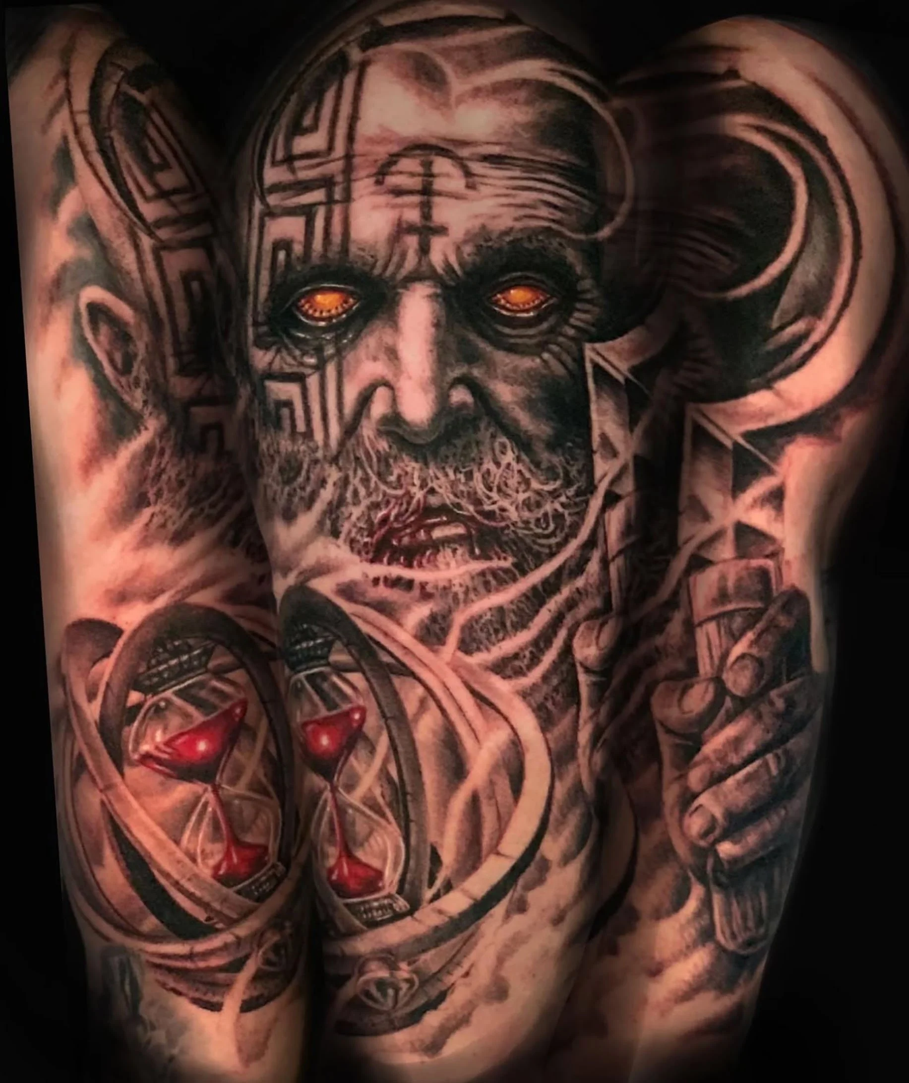 Close-up of a tattooed person's chest and arm featuring a detailed black and gray sculpture of a bearded man's face, with bright orange eyes and a heart-shaped symbol on the chest.