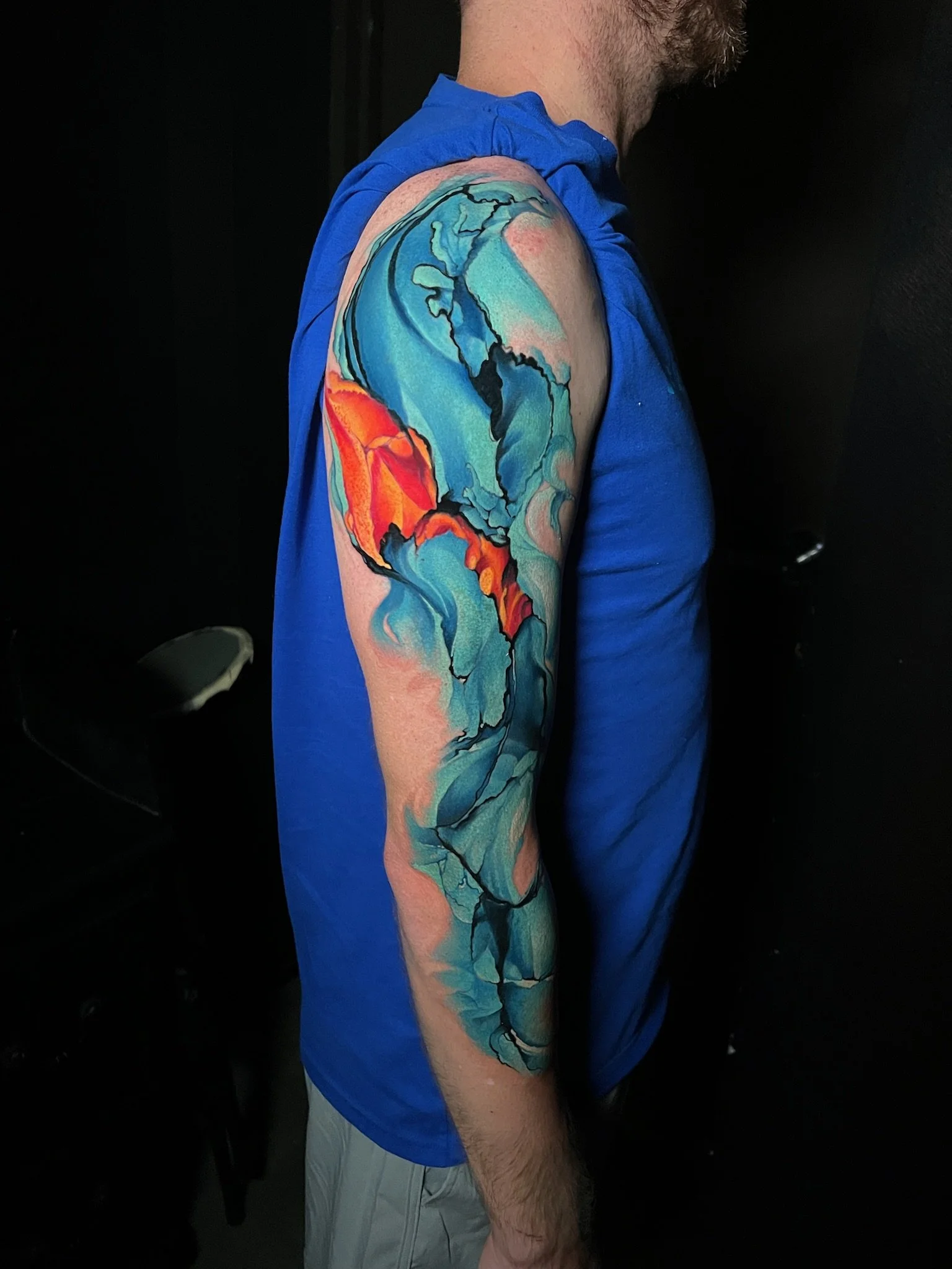 A man with a colorful watercolor style tattoo of a globe on his upper arm and shoulder, wearing a blue sleeveless shirt.