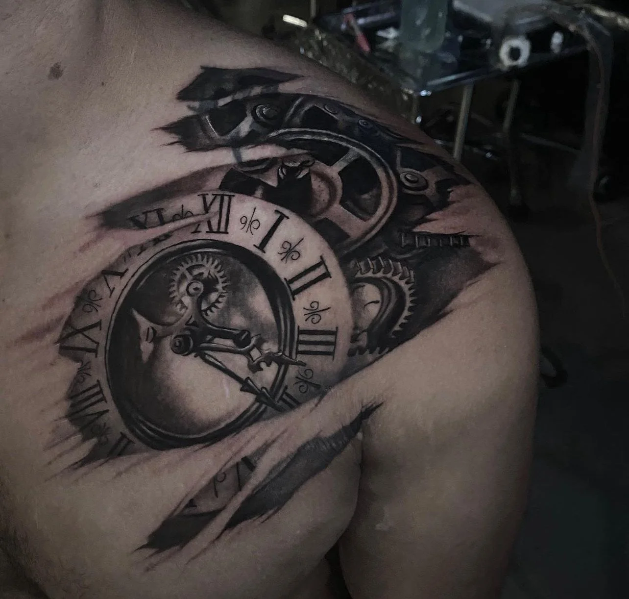A detailed black and gray tattoo of a pocket watch with Roman numerals, gears, and mechanical elements on a person's shoulder and upper arm.