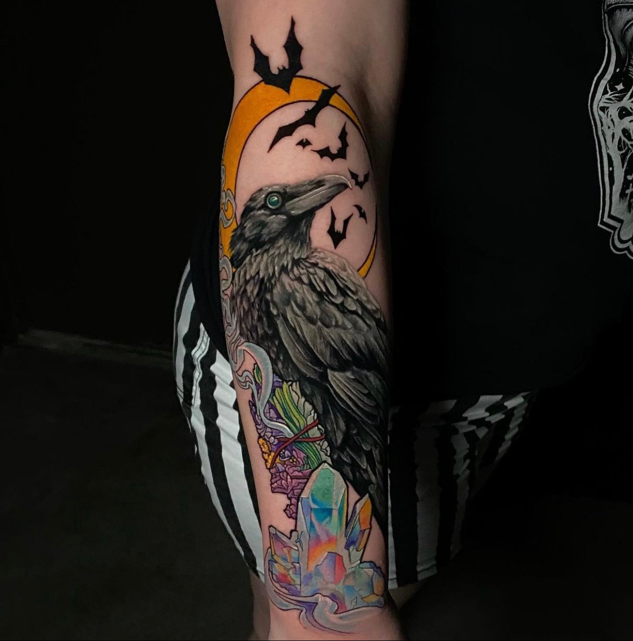 Colorful tattoo of a raven, surrounded by a yellow moon, bats, and a crystal, on a person's arm.