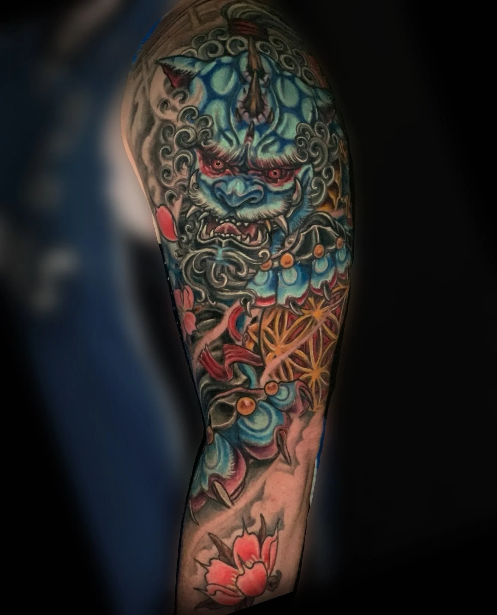 Colorful tattoo on a person's upper arm featuring a fierce blue and white creature with red eyes and sharp teeth, surrounded by clouds, flowers, and other intricate designs.