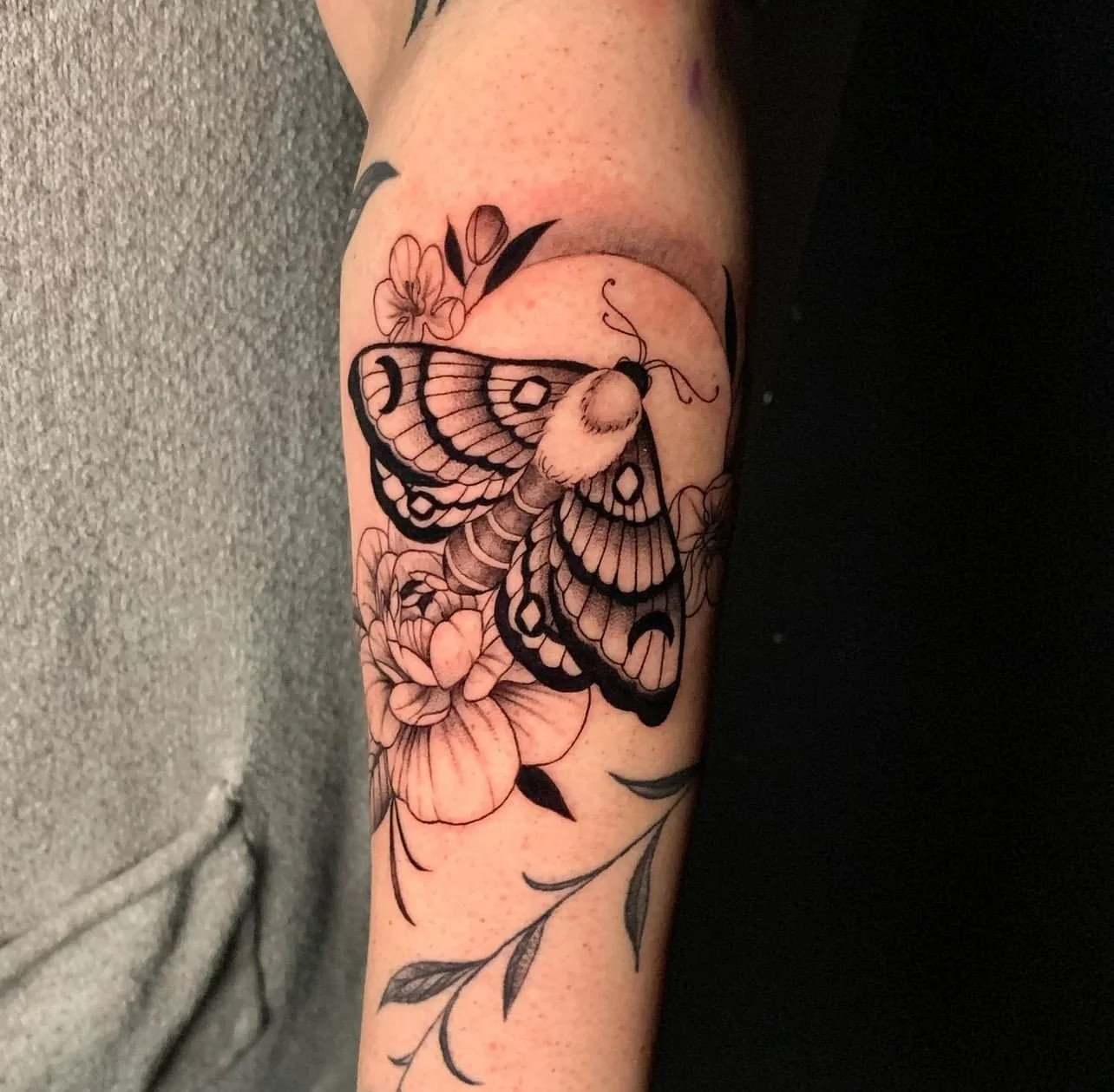 Black and white tattoo of a butterfly perched on a flower with leaves on the arm.