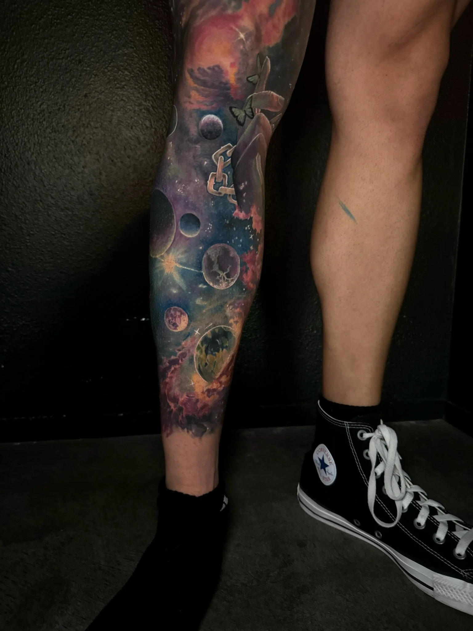 A colorful space-themed tattoo sleeve on a person's leg, depicting planets, moons, stars, nebulae, and celestial elements.