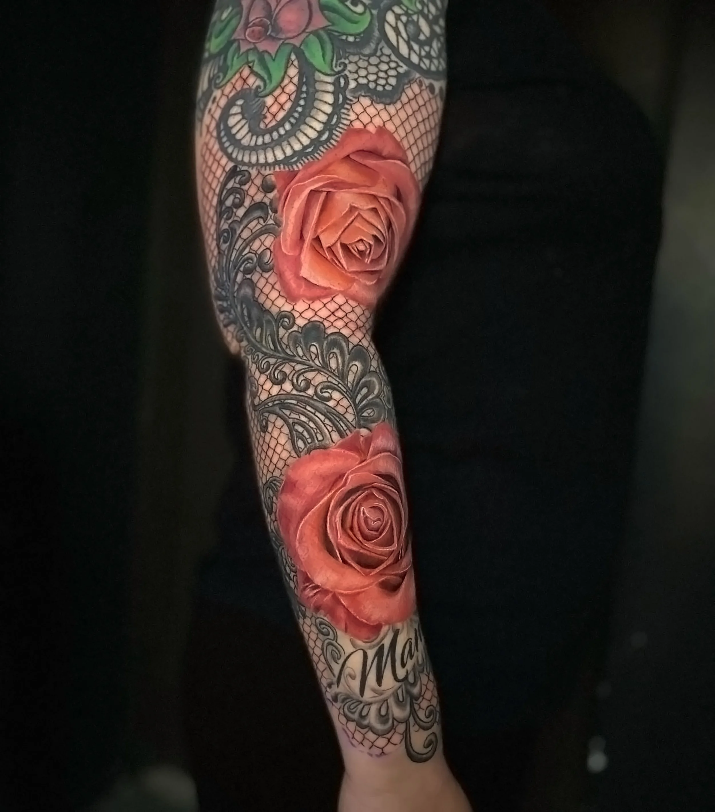 A person's arm with multiple tattoos, including a detailed pink rose with layered petals and black outlines, and intricate designs with floral motifs, leaves, and swirling patterns.