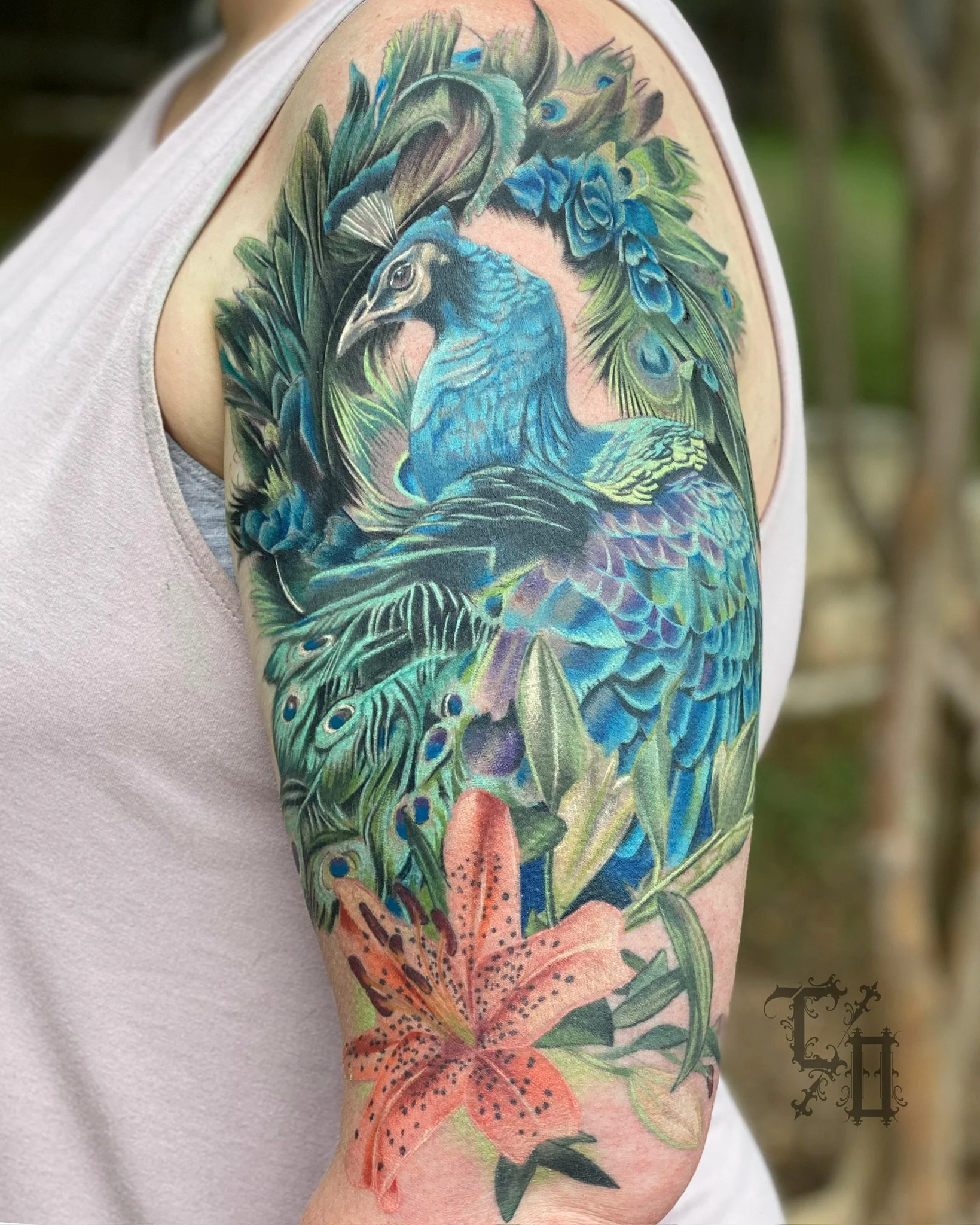Colorful tattoo of a peacock surrounded by leaves and a lily flower on a person's upper arm.