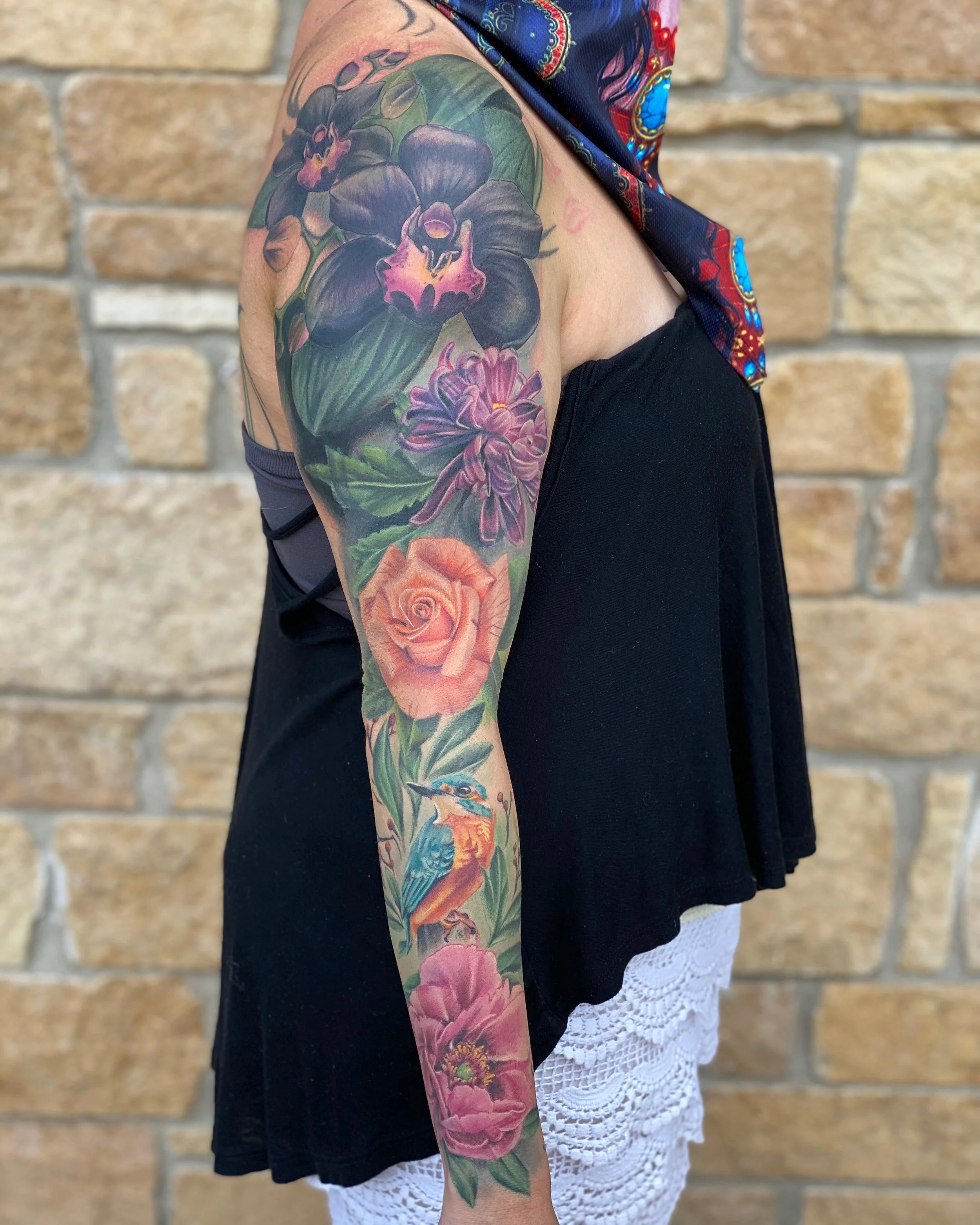 A person's arm with a colorful floral tattoo featuring large dark purple orchids, pink lilies, and a kingfisher bird against a brick wall background.