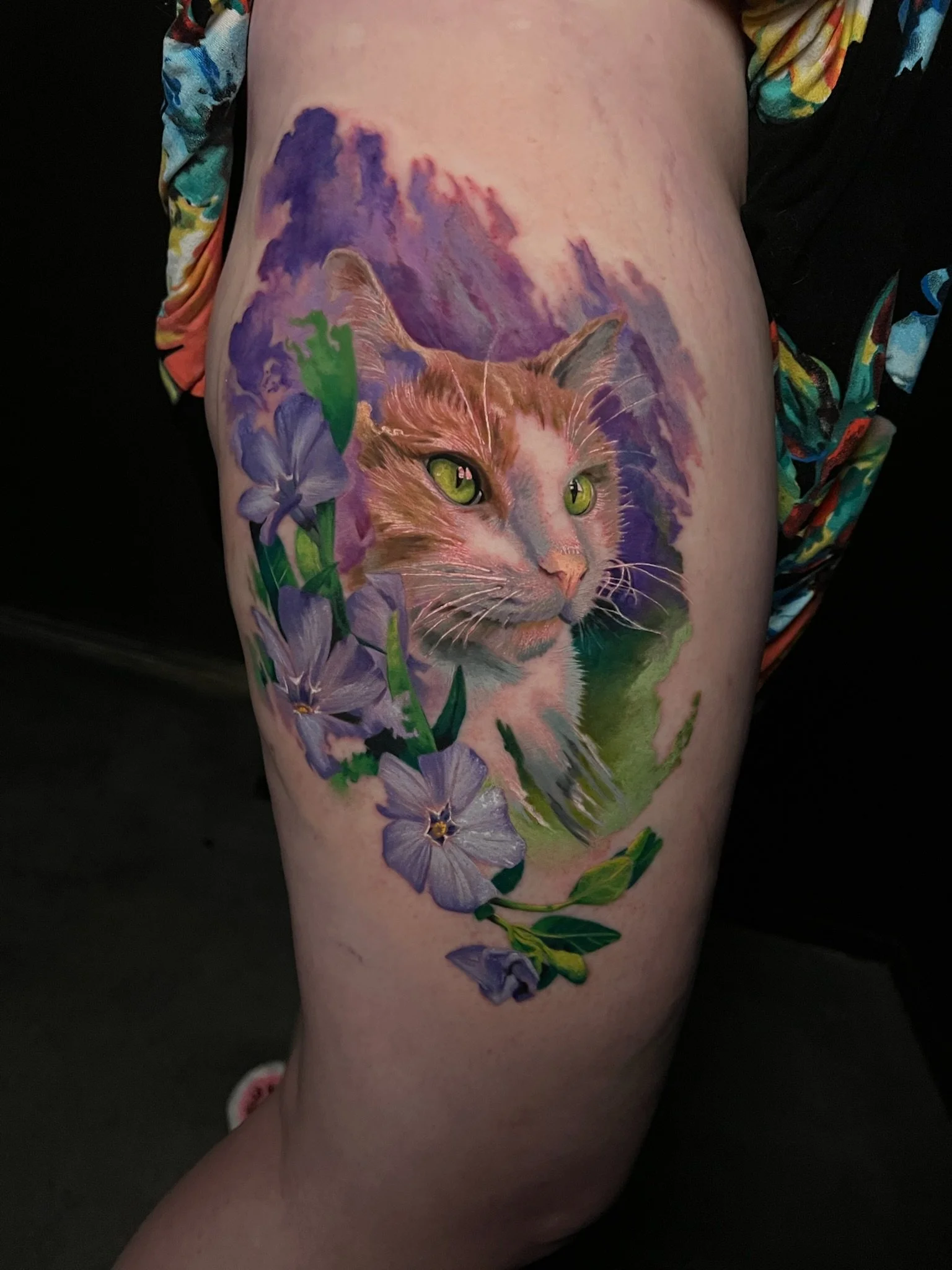 Colorful tattoo of a realistic cat's face with green eyes, surrounded by purple flowers and green leaves, on a person's thigh.