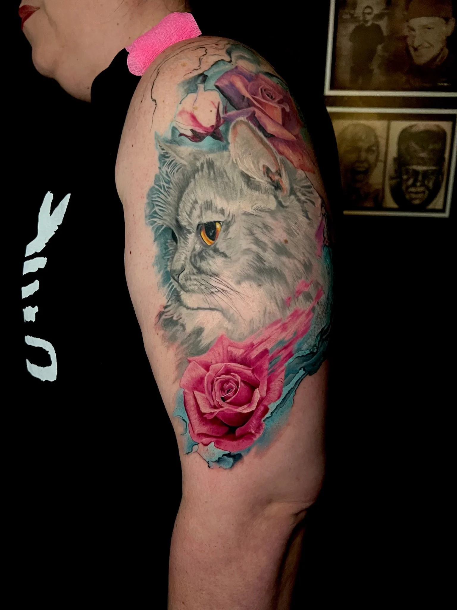 Tattoo of a gray cat's face surrounded by pink roses on a person's upper arm.