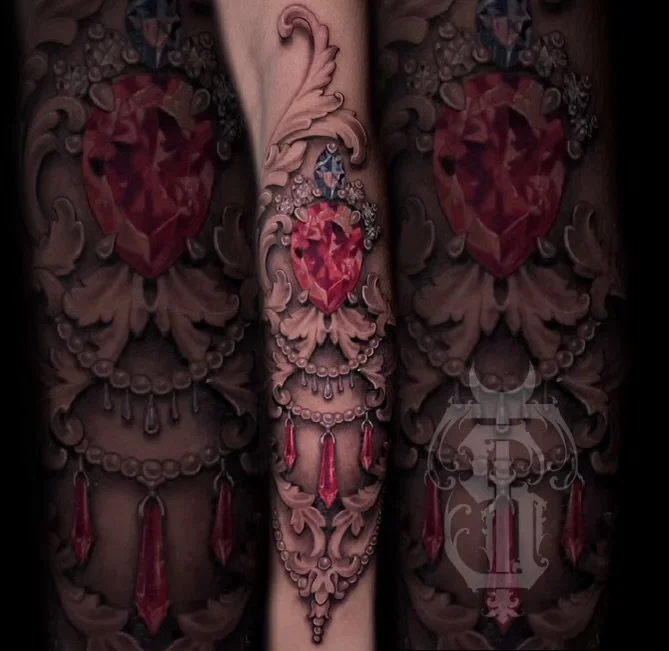 Tattoo of a red heart with red gemstones and jewelry designs on forearm.