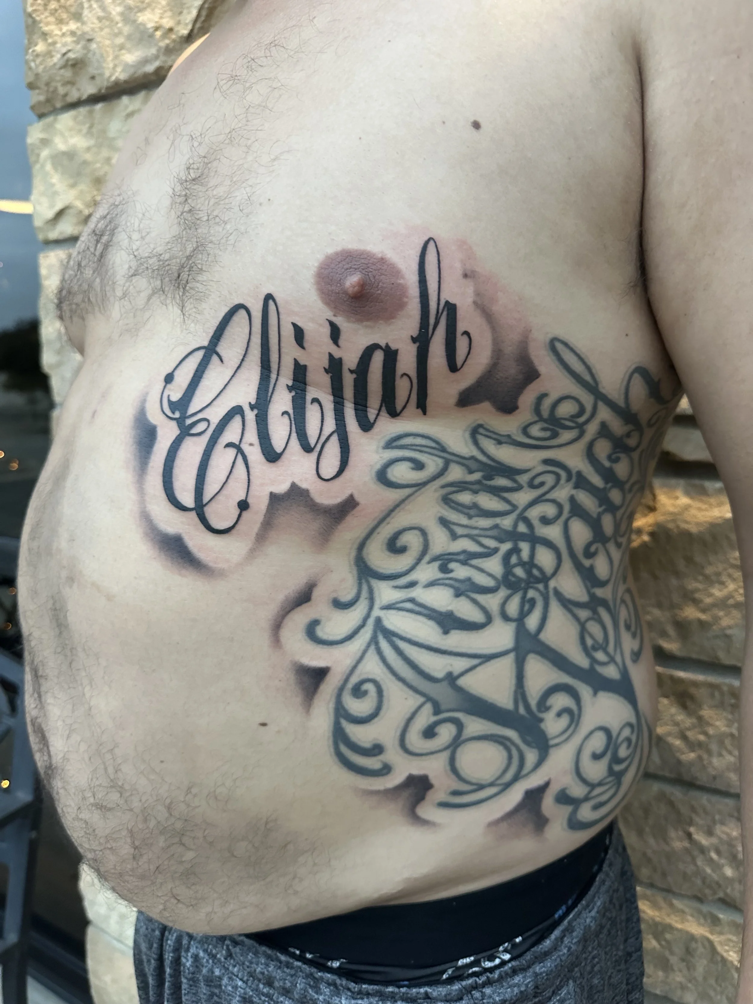 Close-up of a man's upper chest with large black tattoos including the name "Elijah" and ornate swirling designs, with the man wearing dark shorts and standing near a stone wall.