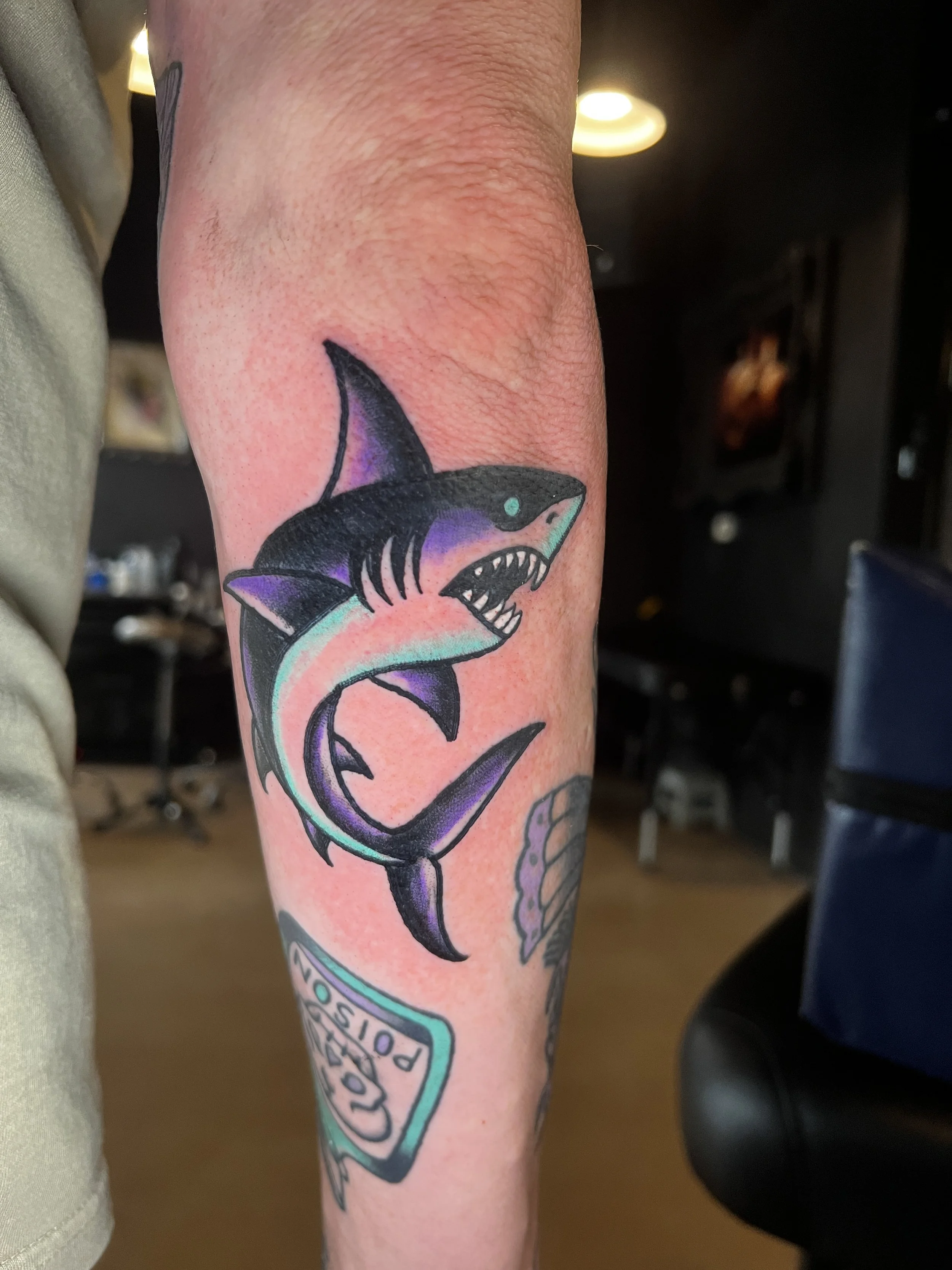 A tattoo of a great white shark on a person's arm, showing the shark's open mouth with sharp teeth and a detailed eye, with a colorful background and some text below.