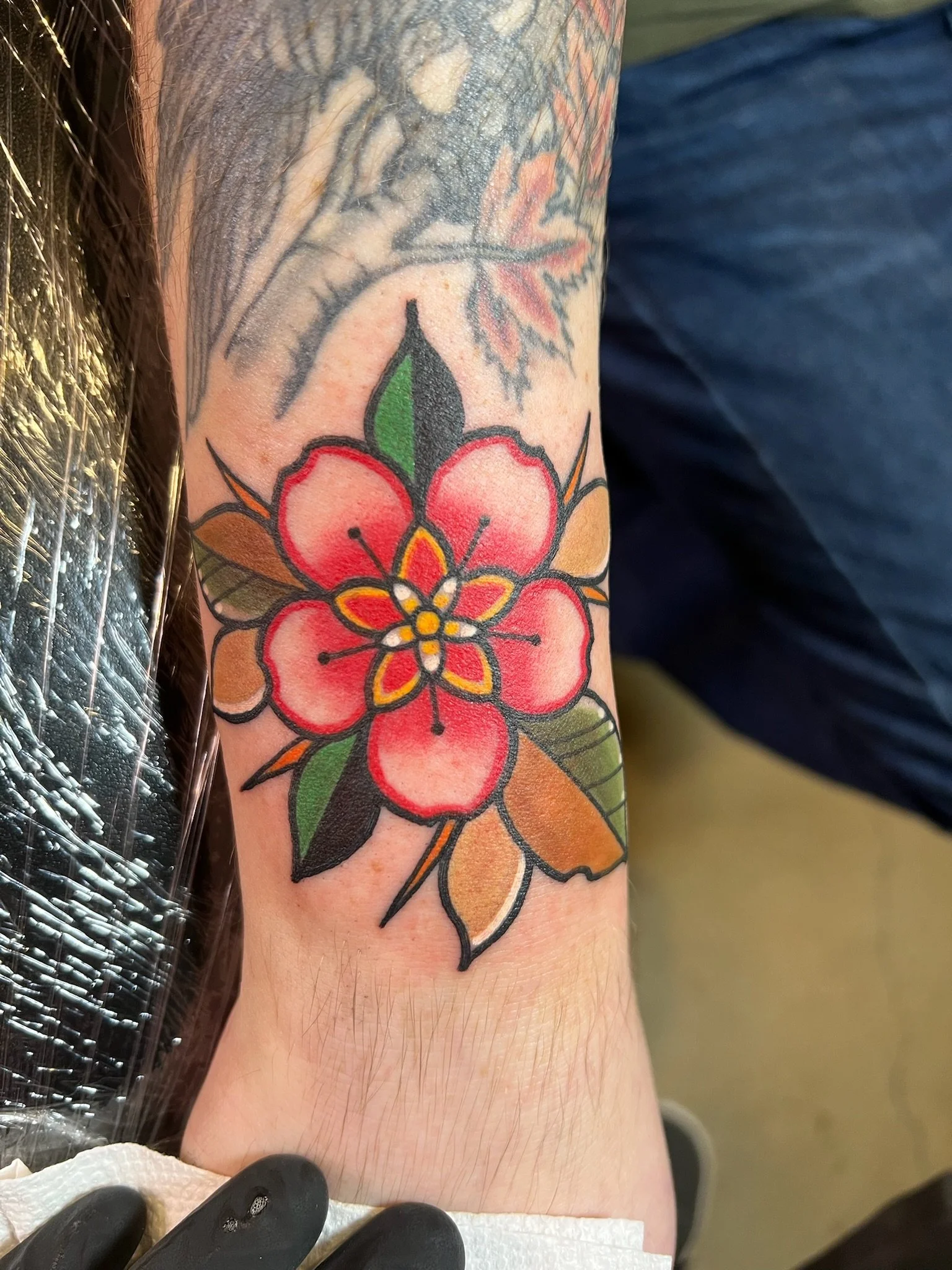 Colorful tattoo of a pink and red flower with green, black, and brown leaves on a person's arm.