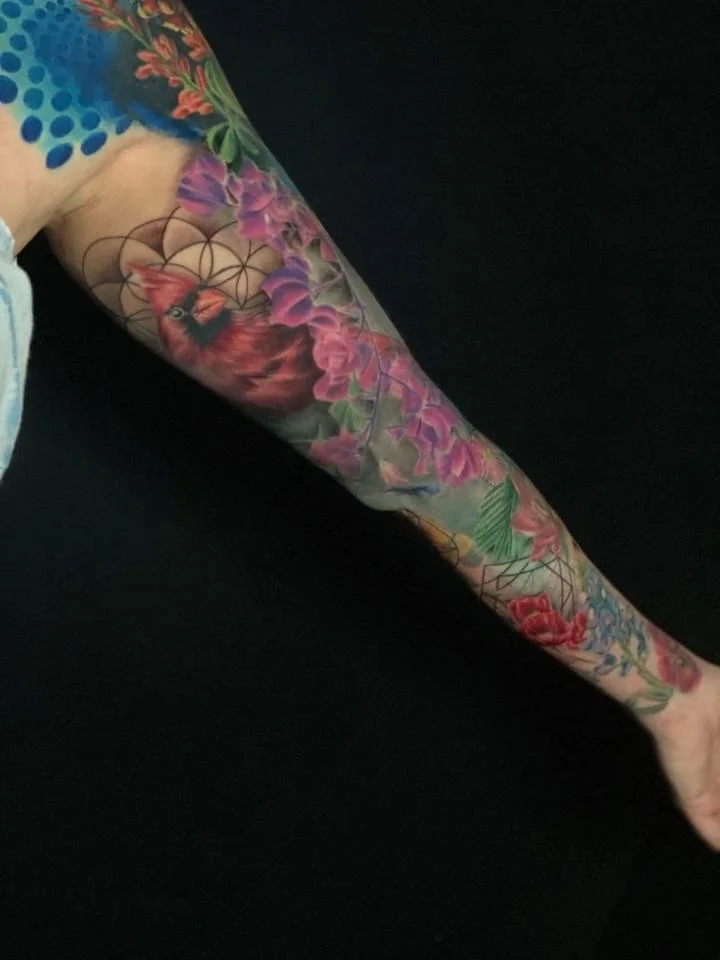 Colorful arm tattoo featuring a bird, flowers, and geometric patterns.