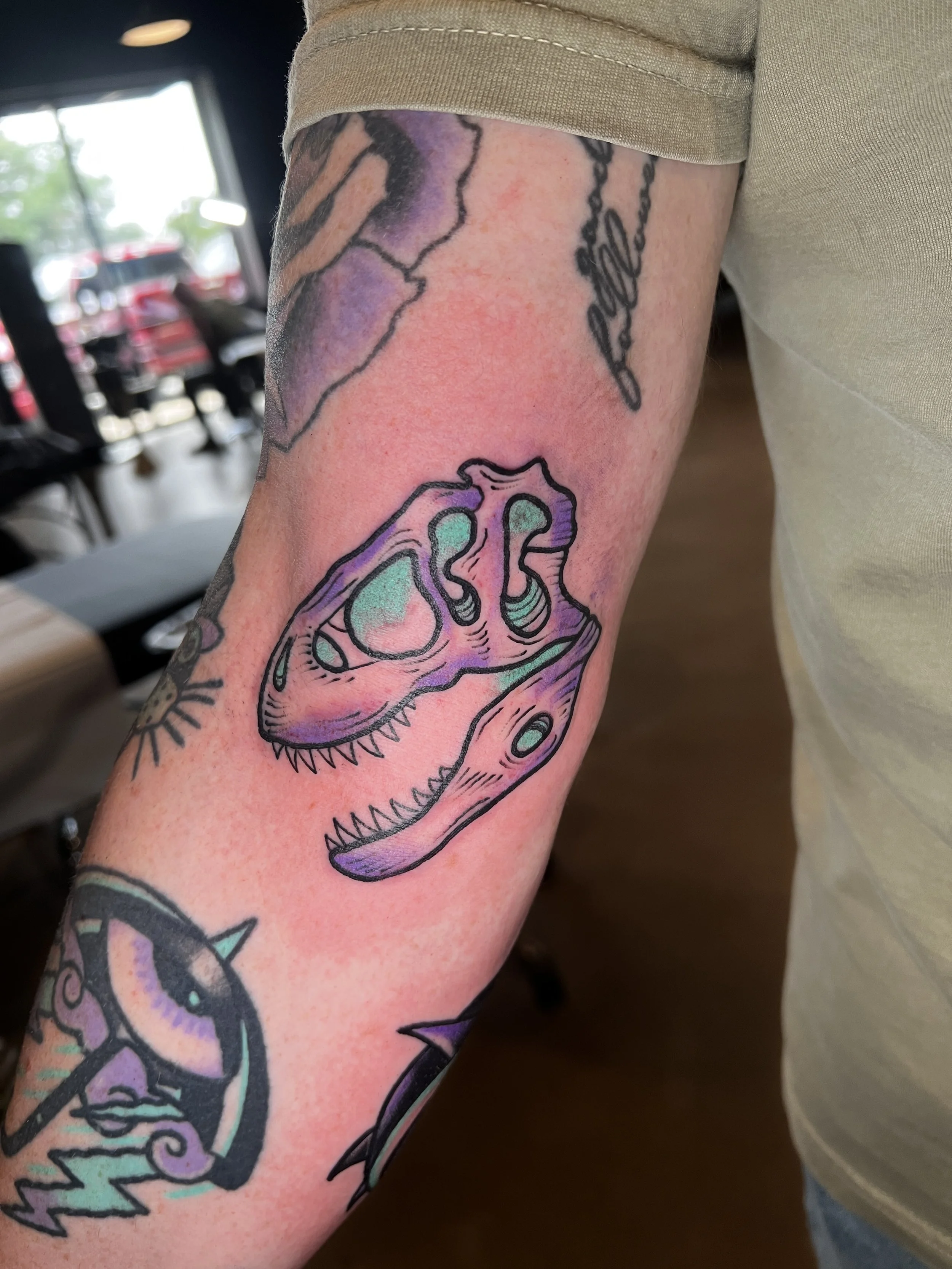 Close-up of a colorful tattoo of a dinosaur skull with green eyes on a person's arm.