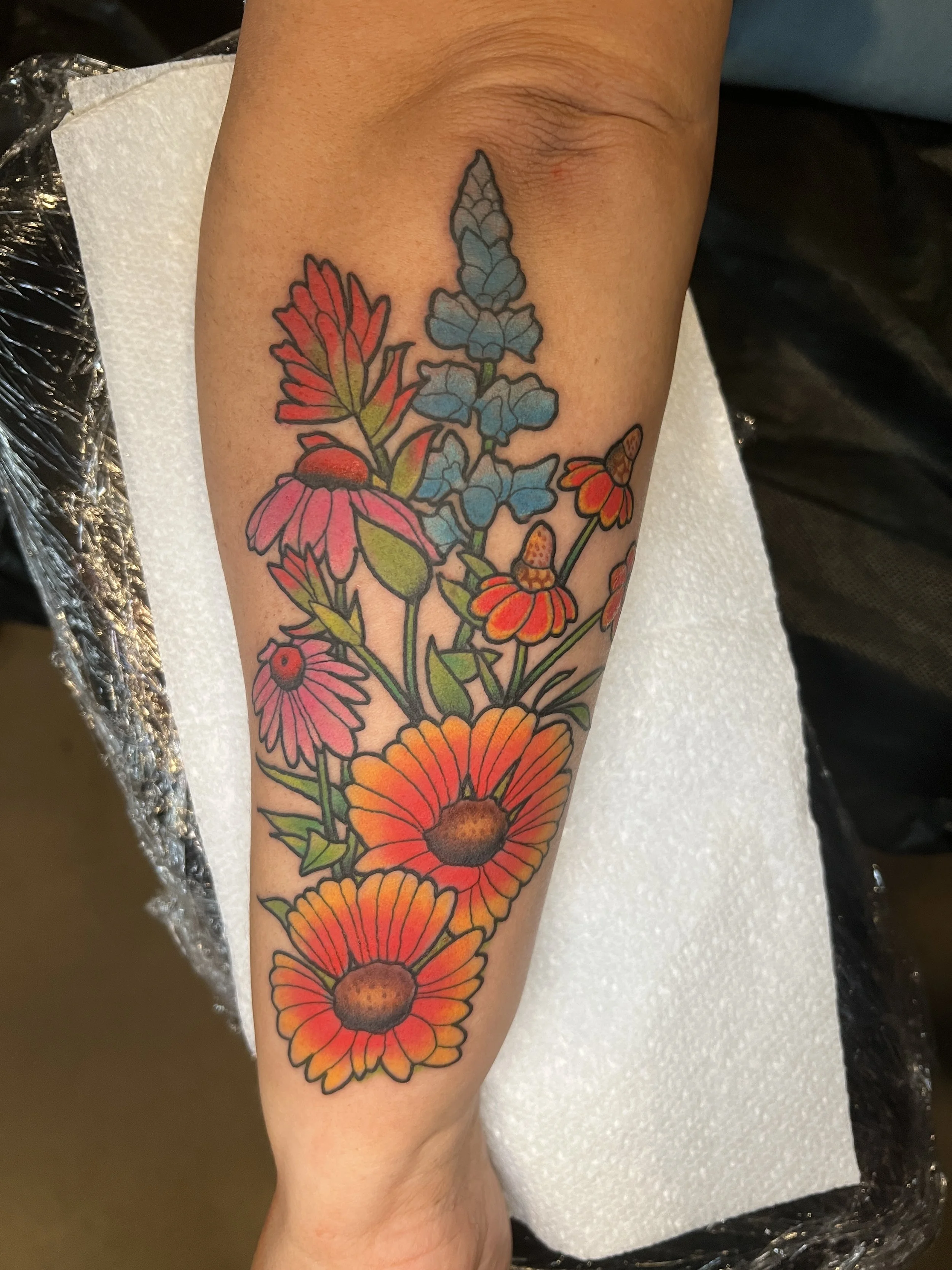 colorful tattoo of a purple, blue, orange, and pink bouquet of flowers on the outside of a someone's forearm