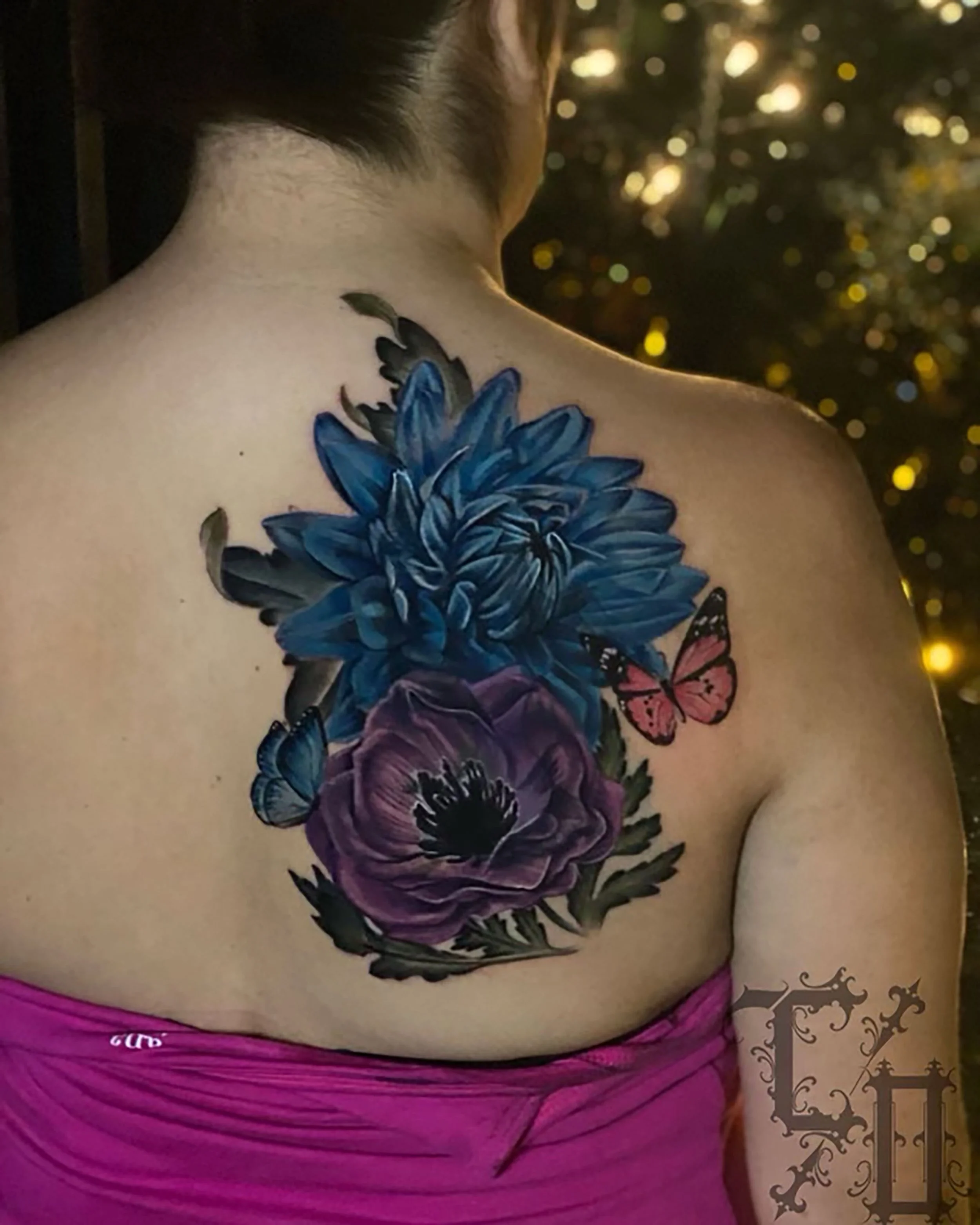 A person with a large floral tattoo on their upper back, featuring blue, purple, and black flowers and two butterflies, wearing a pink garment, with a blurred background of sparkling lights.