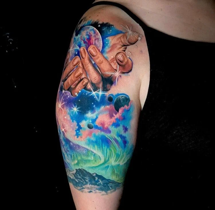 Colorful tattoo of two hands touching with cosmic and mountain scene details.