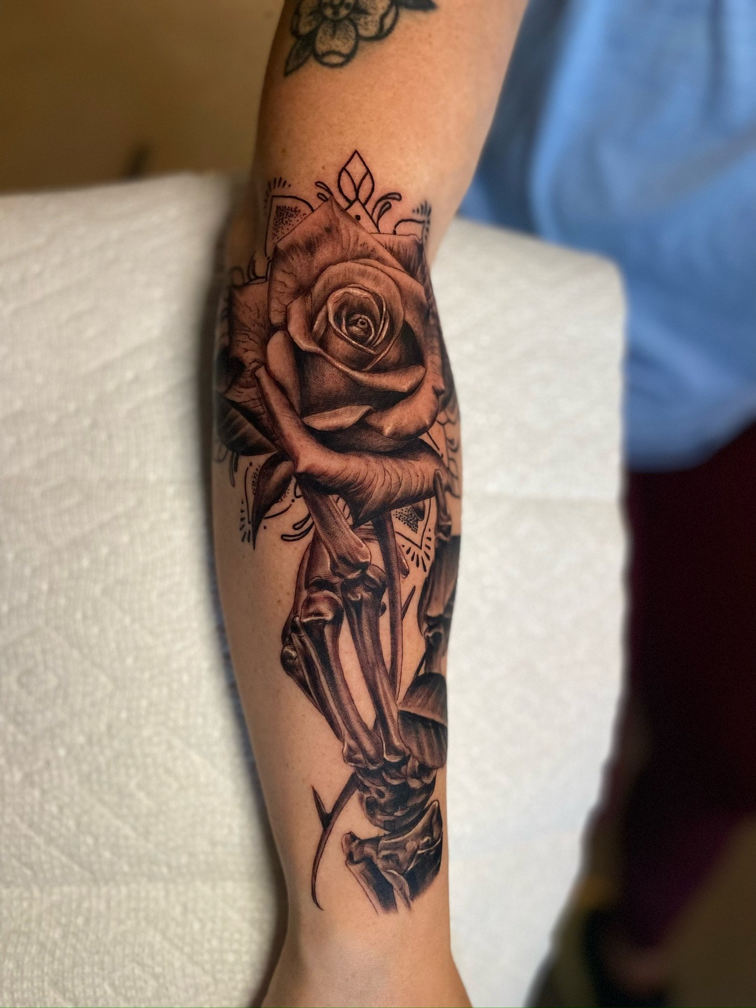 Close-up of a person's arm with a detailed black and gray tattoo of a rose with petals and leaves, combined with skeletal bones below it.