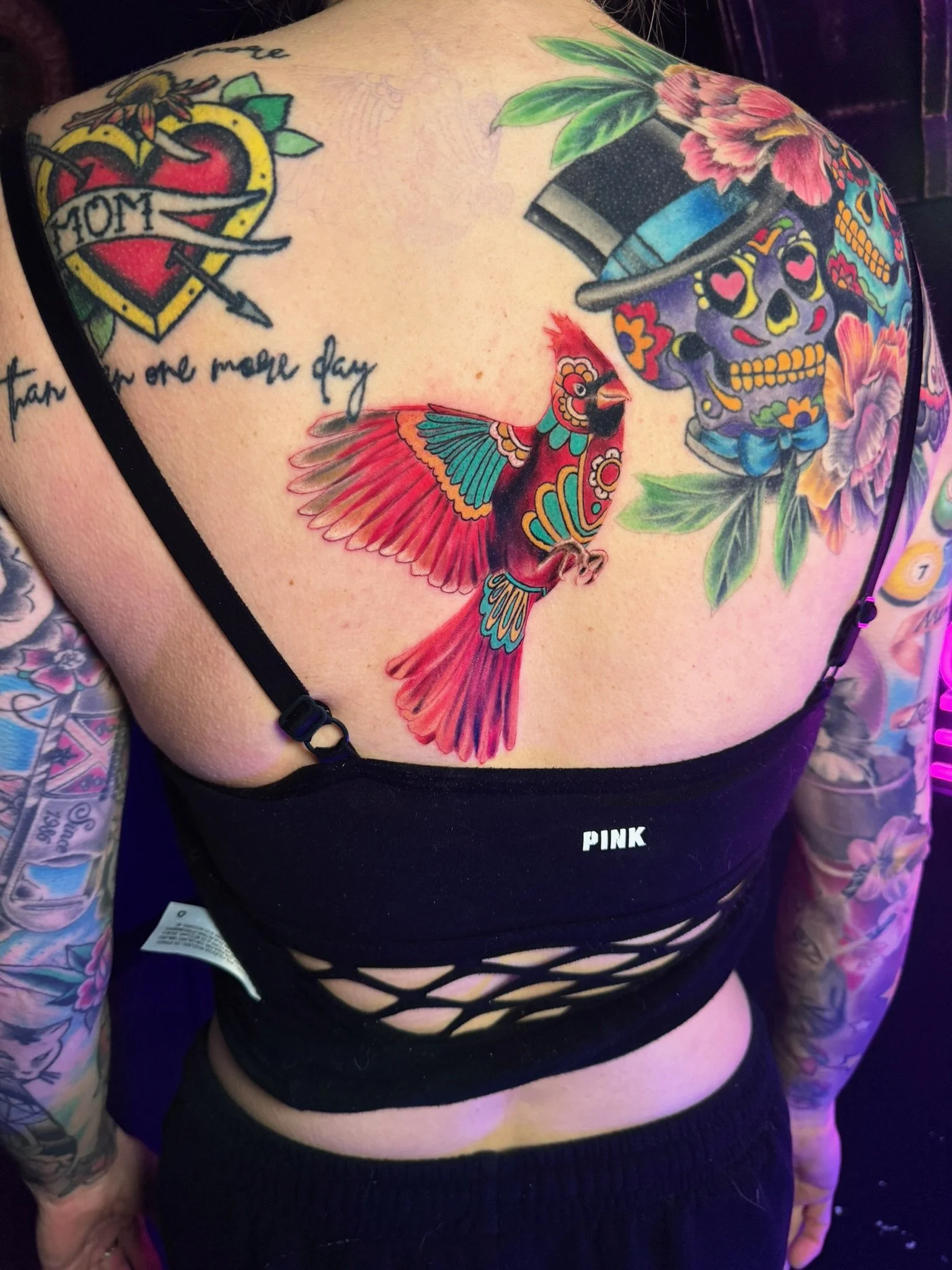 Colorful tattoos on a woman's chest and arms, including a sugar skull with a top hat, a red and turquoise bird, a heart with a banner that reads 'MOM,' and various floral designs.