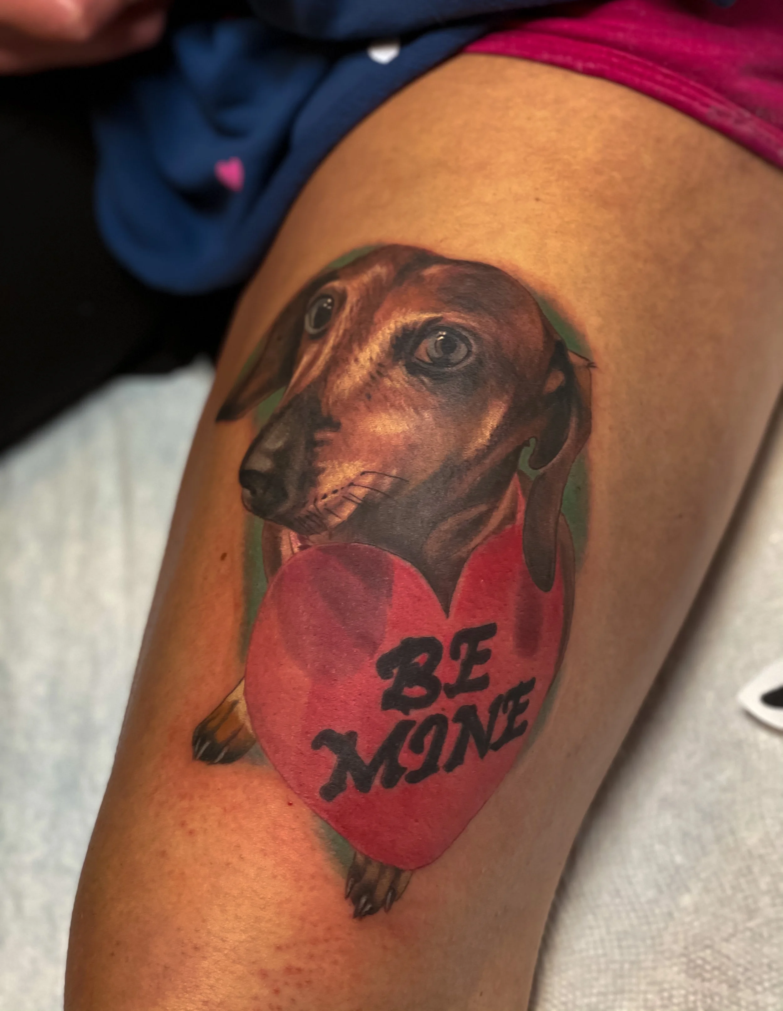 Tattoo of a realistic dog with brown and black fur, large eyes, and a red collar, on a person's arm with a red heart-shaped background and the phrase 'BE HONEST' written in black letters.