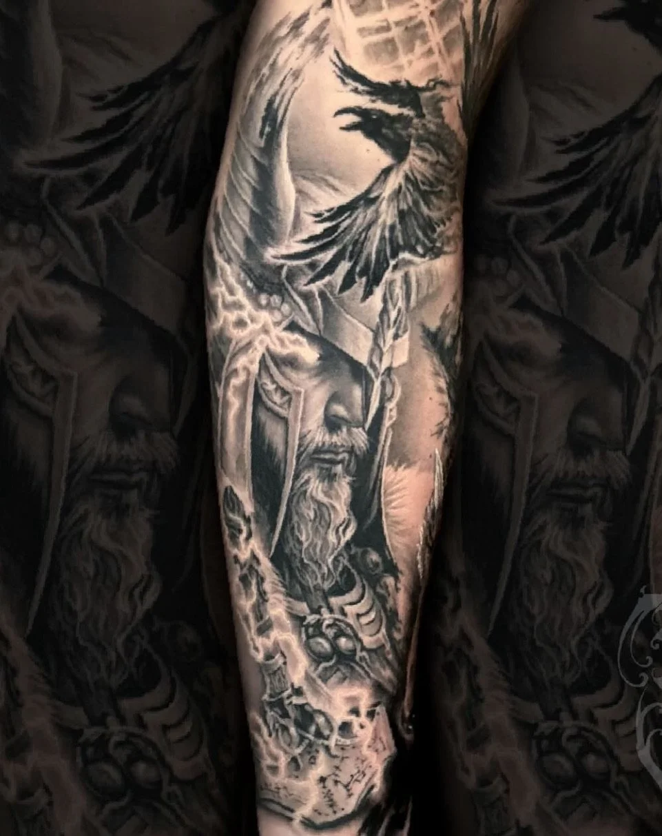 Black and gray tattoo of a bearded man wearing a helmet with a feathered or winged crest, and holding a shield, depicted on a person's arm.