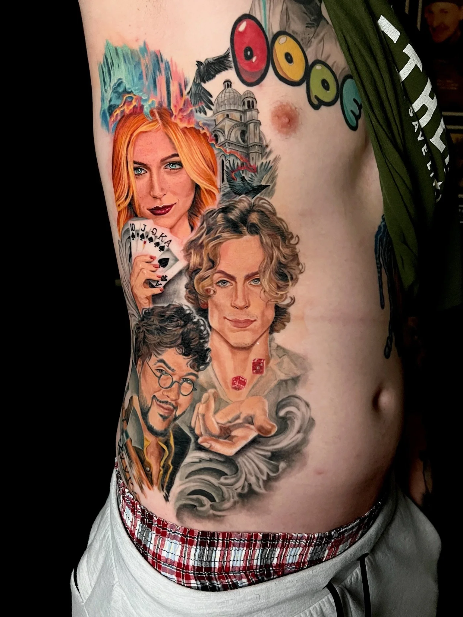 Colorful tattoo of three women with detailed facial features, a hand holding playing cards, playing dice, a building, birds, and a "DOTO" sign on the person's chest.