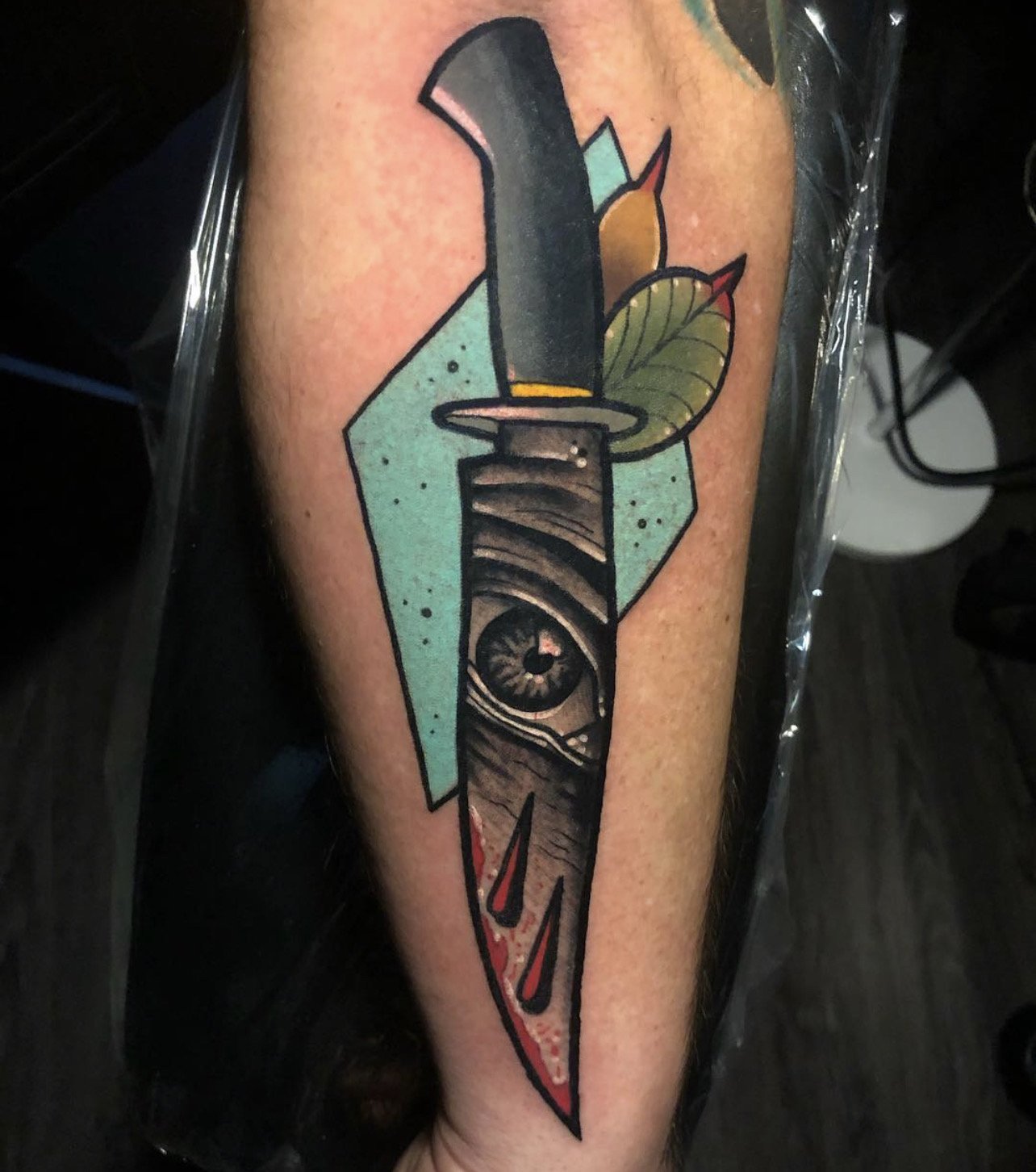 a colorful tattoo of a knife on a person's outer forearm with an eye inside the blade and blood dripping down, with green leaves and a teal abstract shape design behind it