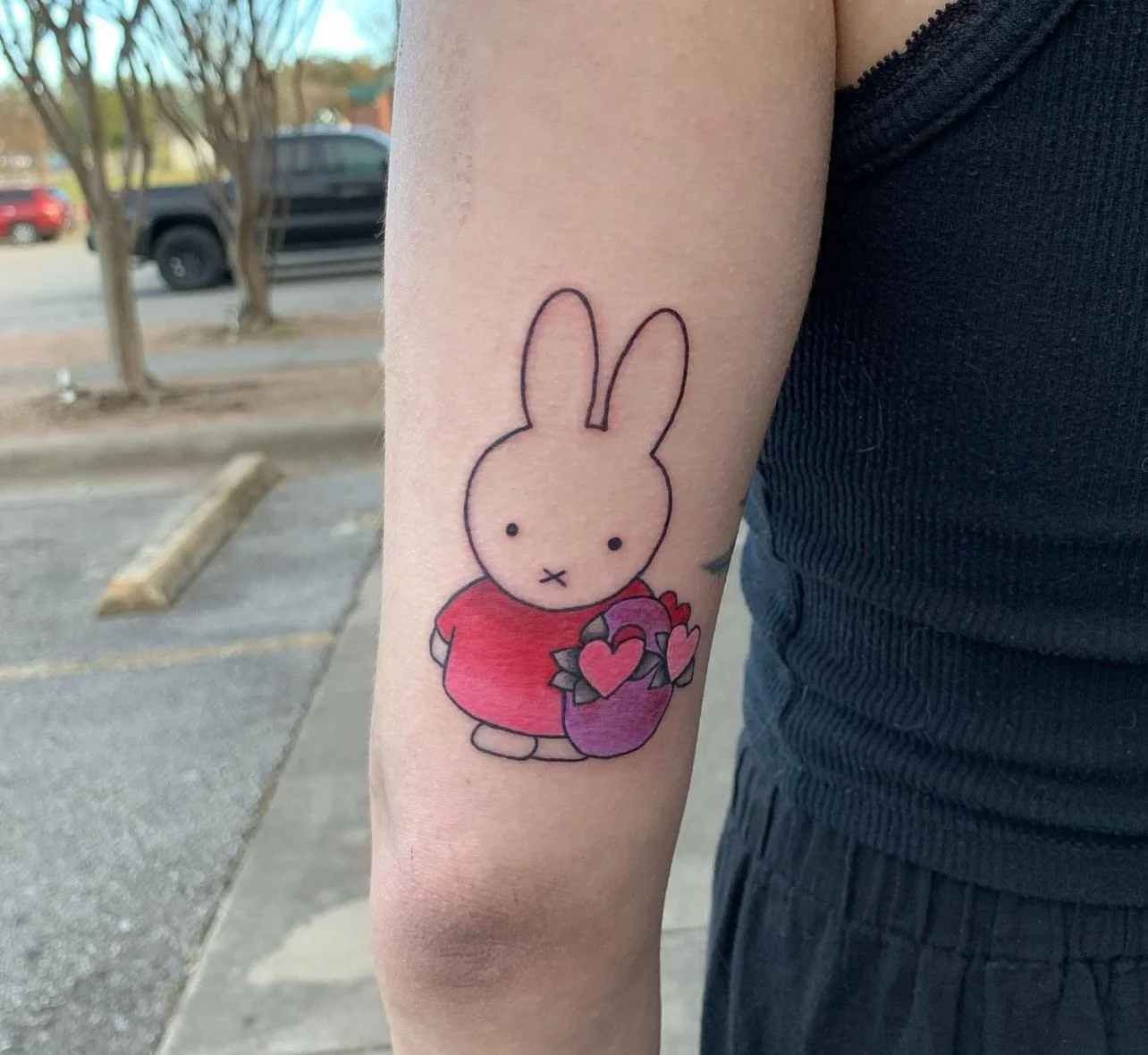 A tattoo of Miffy, a cartoon rabbit, wearing a red shirt and holding a purple and pink flower with heart-shaped petals.