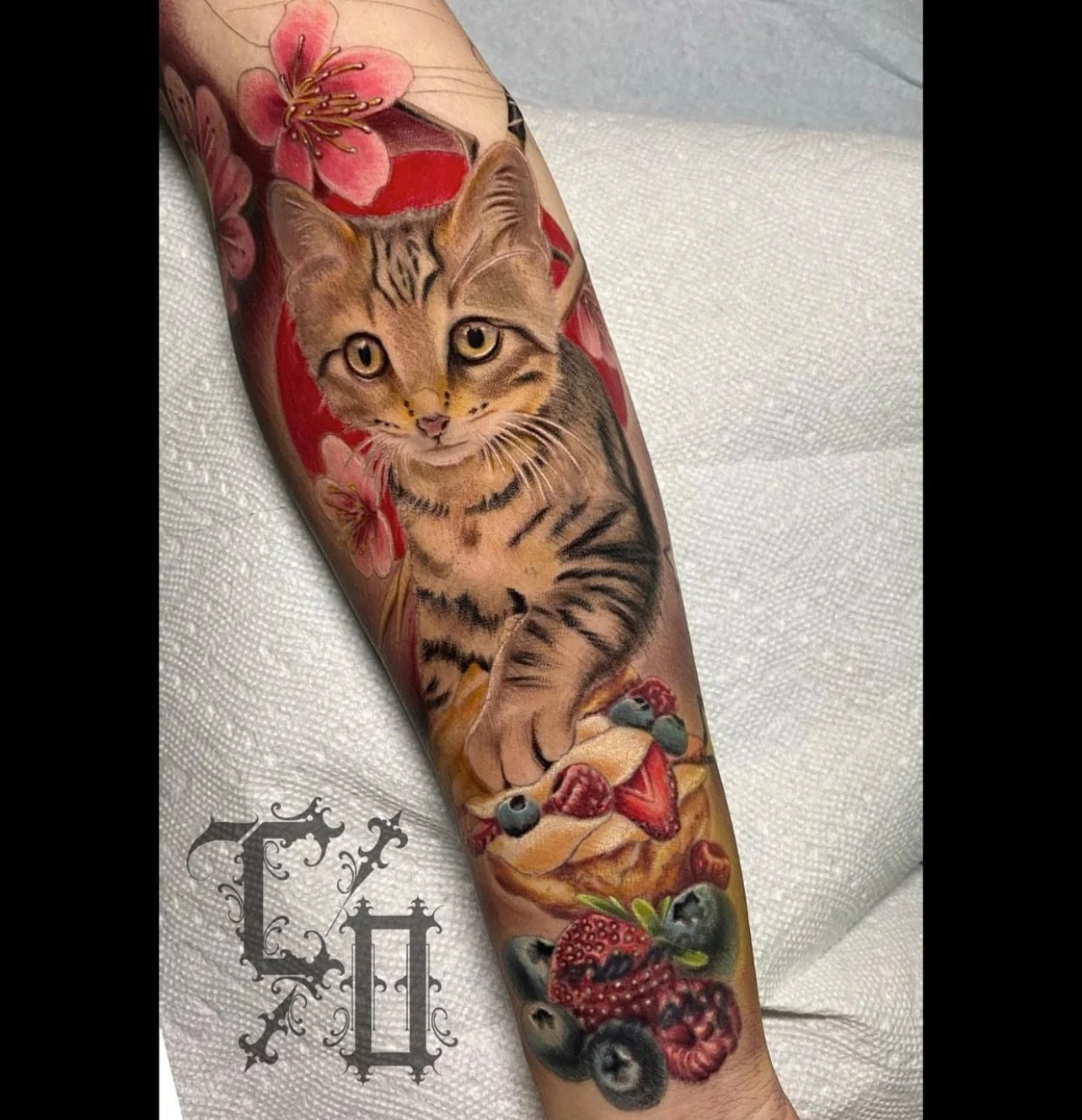 Tattoo of a kitten with a slice of strawberry tart and berries, surrounded by pink flowers, on a person's arm.