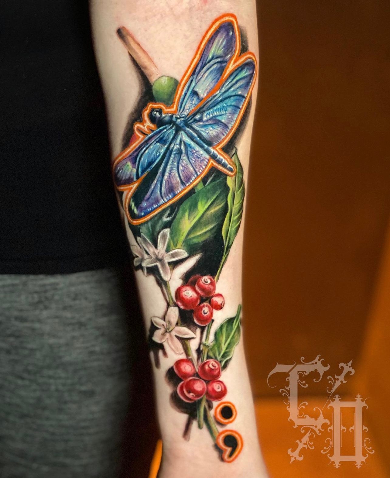 Colorful tattoo of a blue dragonfly with orange outline perched on green leaves, with white flowers, red berries, and black and orange accents on a person's forearm.