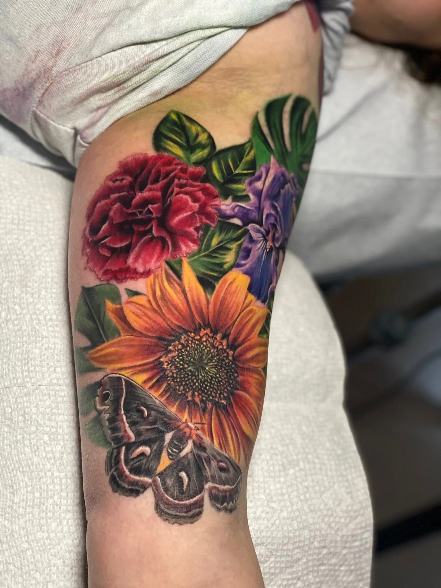 Colorful tattoo of a sunflower, red carnation, blue iris, and butterfly on a person's arm.