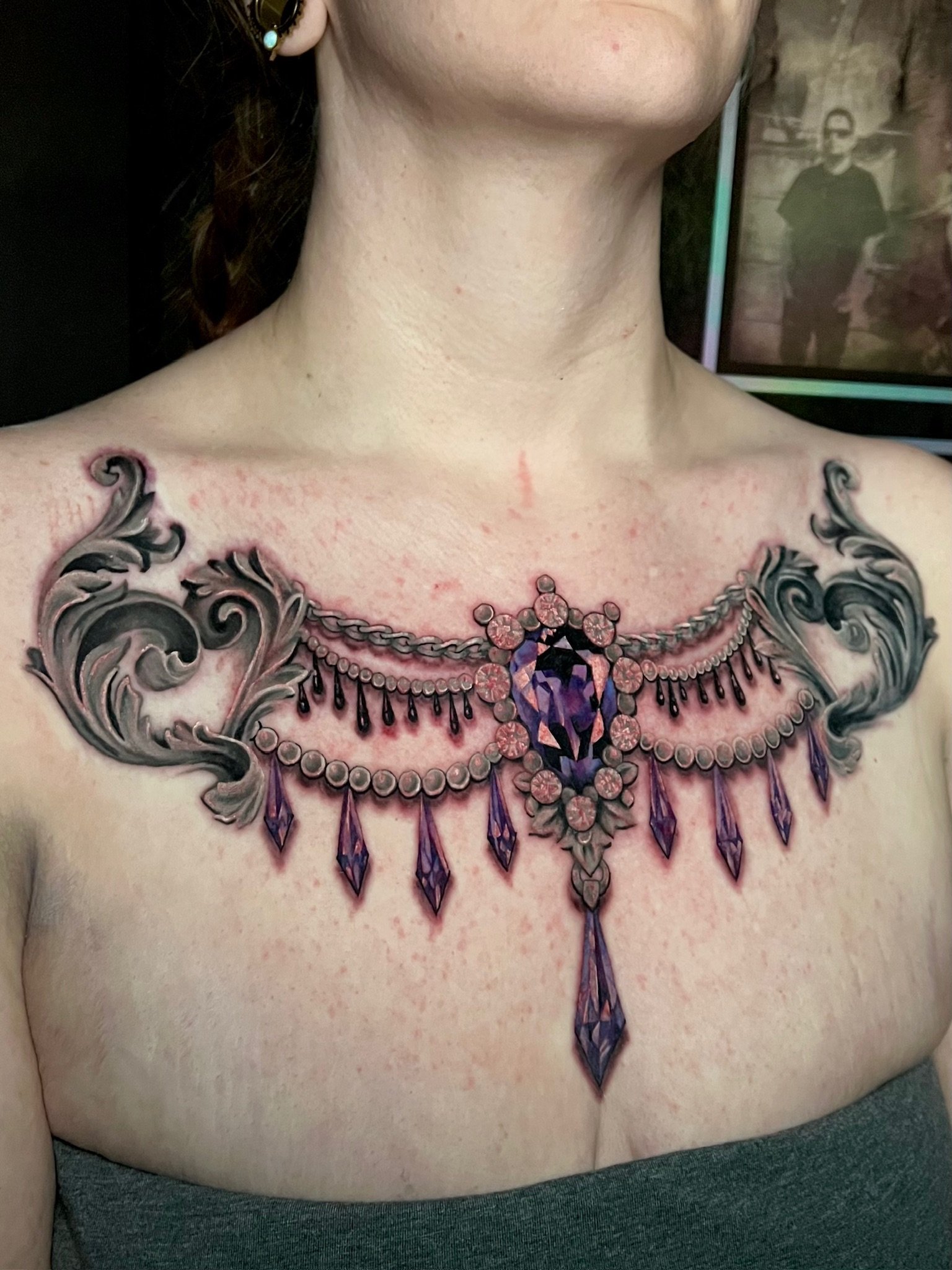 Tattoo of a purple gemstone necklace with black and purple dangling gemstones, ornate scrollwork, and floral details.