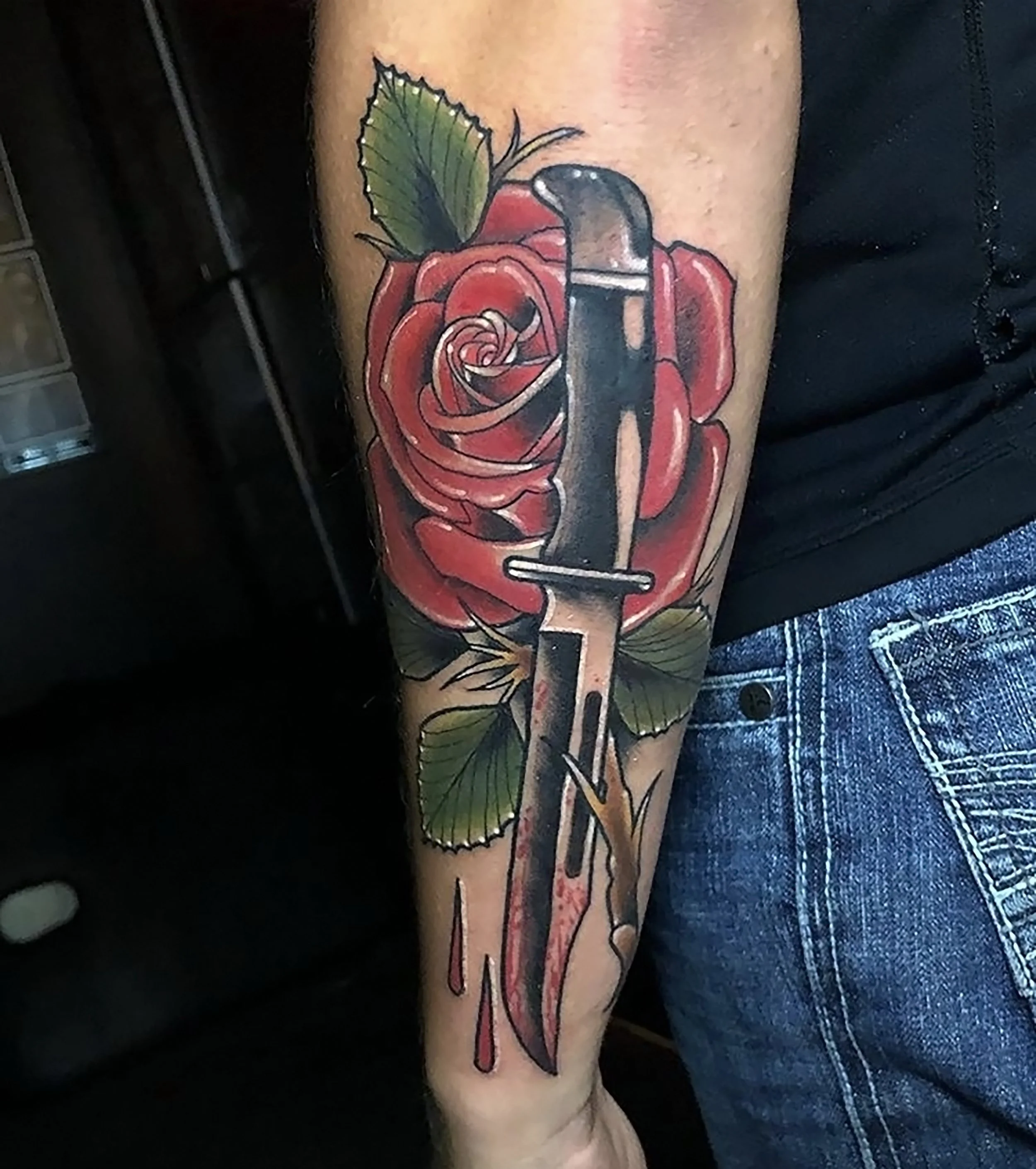 a red, green, and black tattoo of a knife with a red rose, green leaves, and red blood dripping down someone's forearm