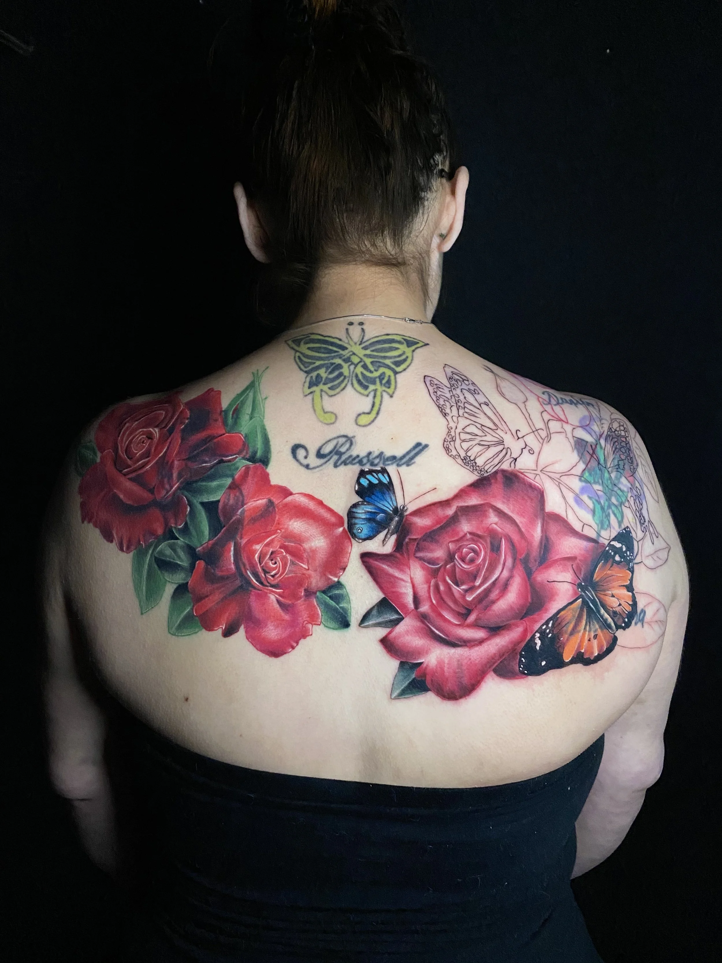 Back of person with tattoos of red roses, butterflies, and the name Russell on the upper back.