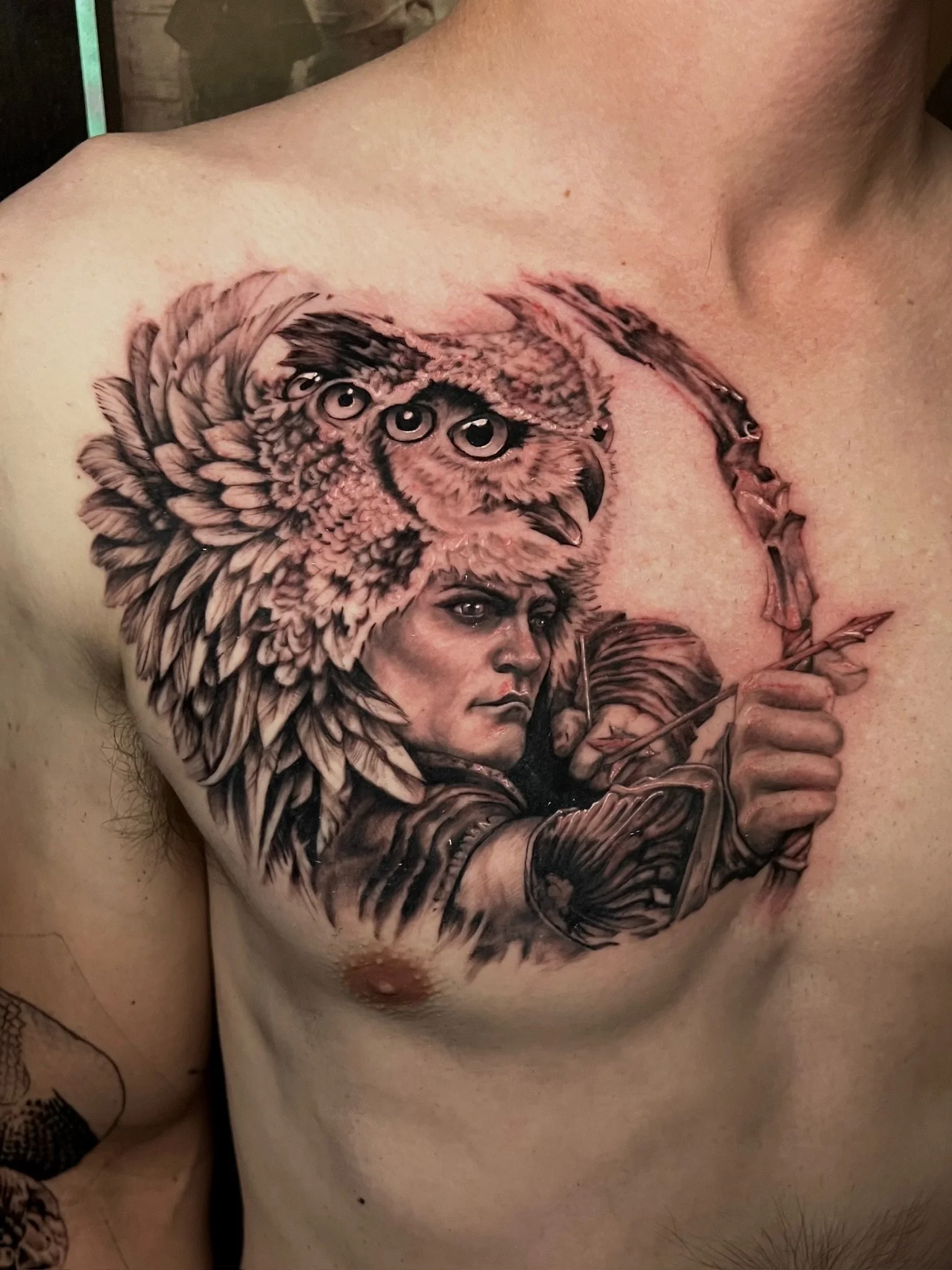 A detailed black and gray tattoo of an owl with three eyes, a human face, and a man holding a bow and arrow, on a person's chest.