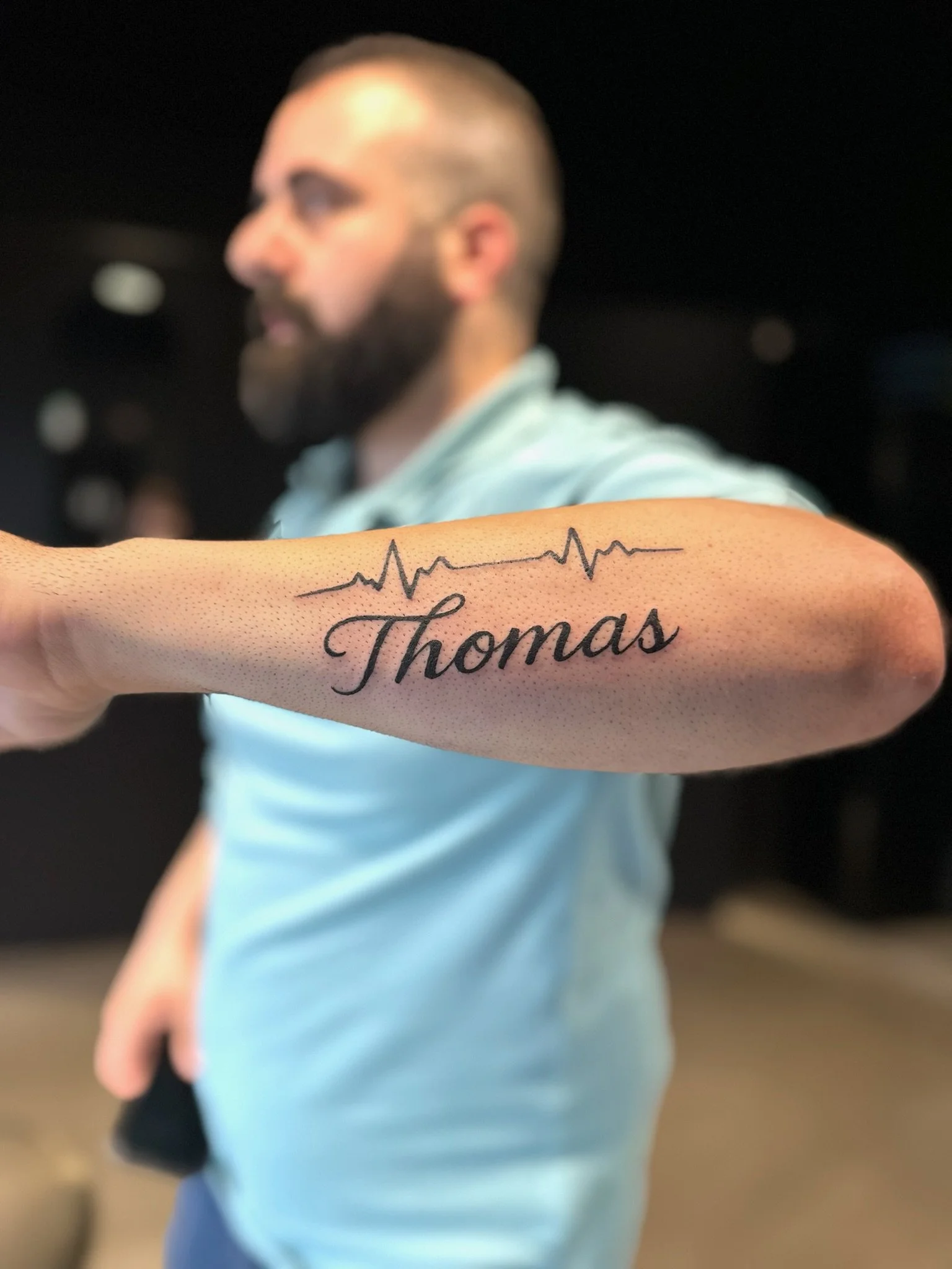 Close-up of a tattoo on a person's forearm that features a heartbeat line above the name 'Thomas' written in cursive. In the background, a man with a beard and short hair is slightly out of focus, wearing a light blue shirt.