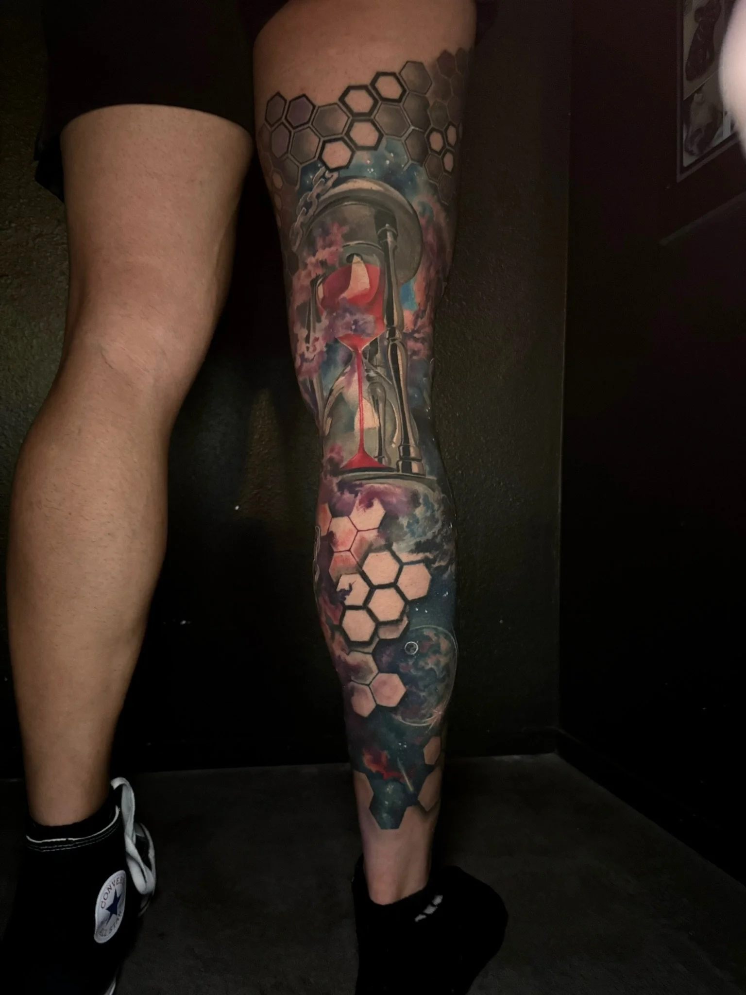 Colorful tattoo of an hourglass with clouds, a galaxy, and hexagons on a person's leg.