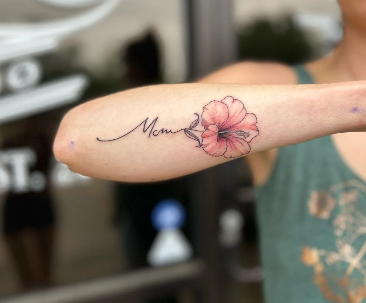 A tattoo on a person's arm featuring the word "Mom" in cursive and a pink hibiscus flower with detailed petals.