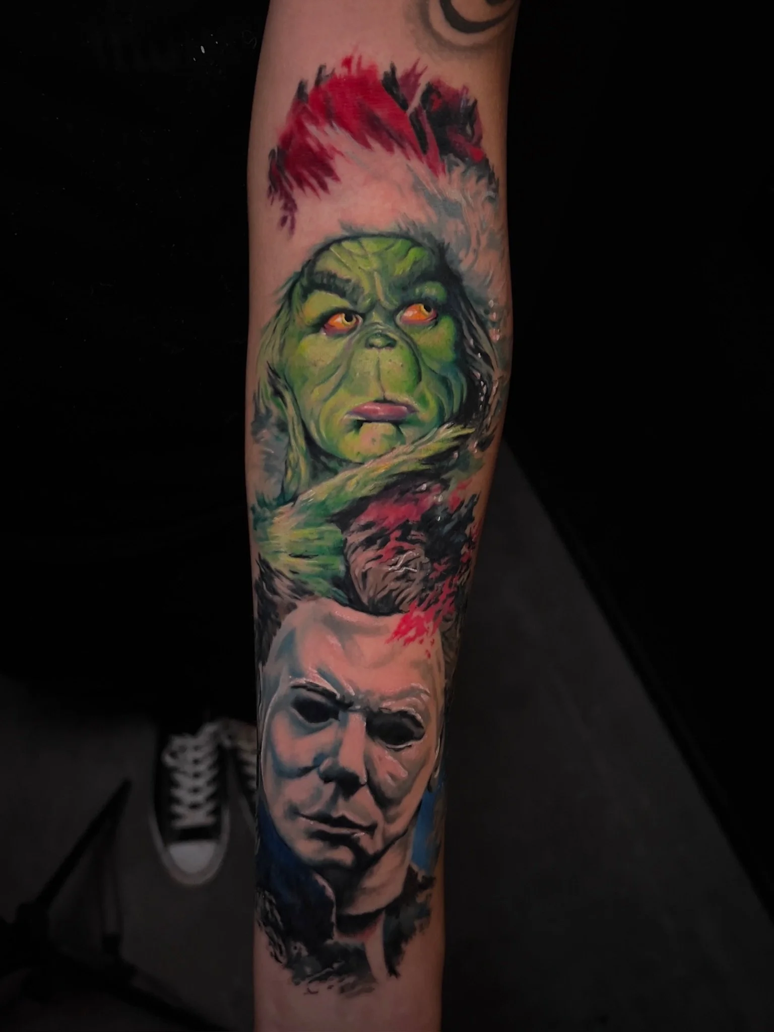 Colorful tattoo of a green creature resembling the Grinch above a realistic portrait of Michael Myers from the Halloween movie series on a person's forearm.