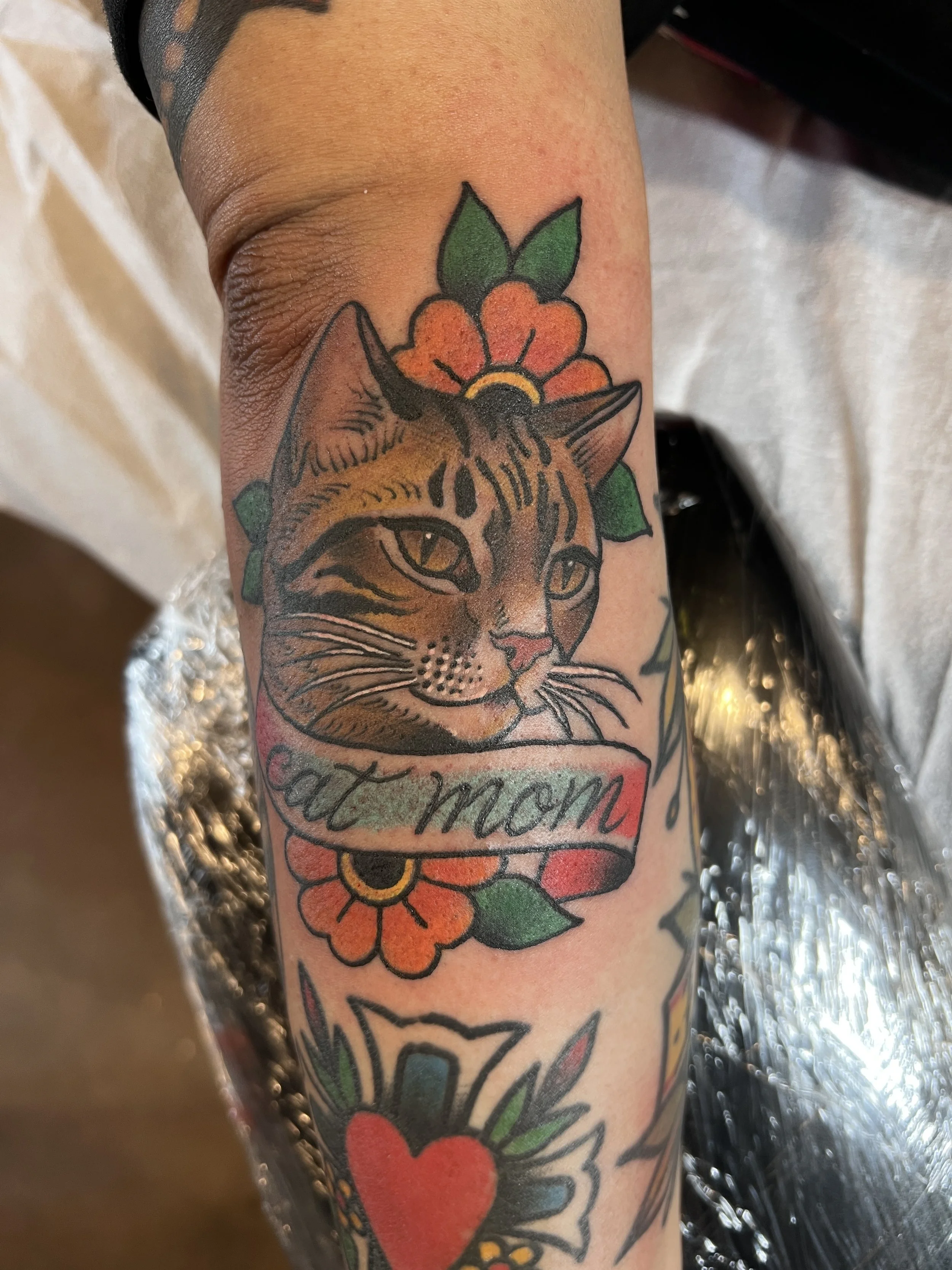 Close-up of a tattoo on a person's arm featuring a cat's face with green leaves and orange-red flowers above its head, a red heart below, and the word 'Mama' in cursive script.