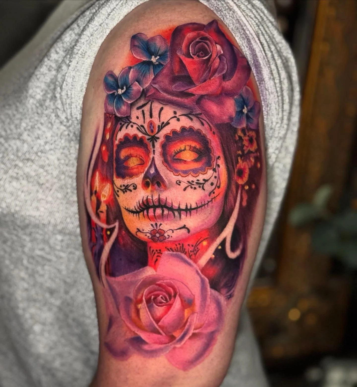 Colorful tattoo of a skull with a sugar skull design, adorned with purple and blue flowers on the forehead, and pink roses at the bottom.