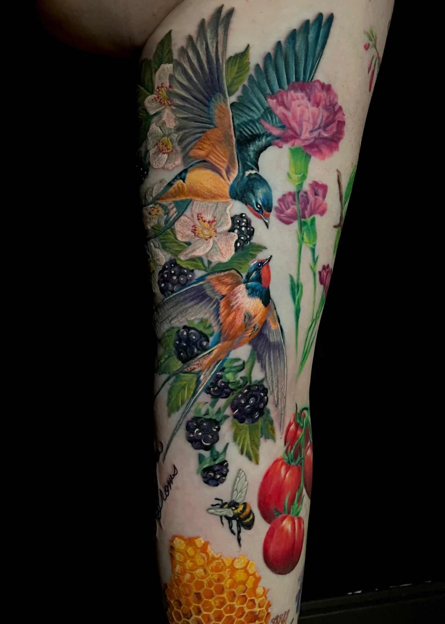 Colorful tattoo of two birds, flowers, blackberries, a bee, honeycomb, and tomatoes on a person's arm.