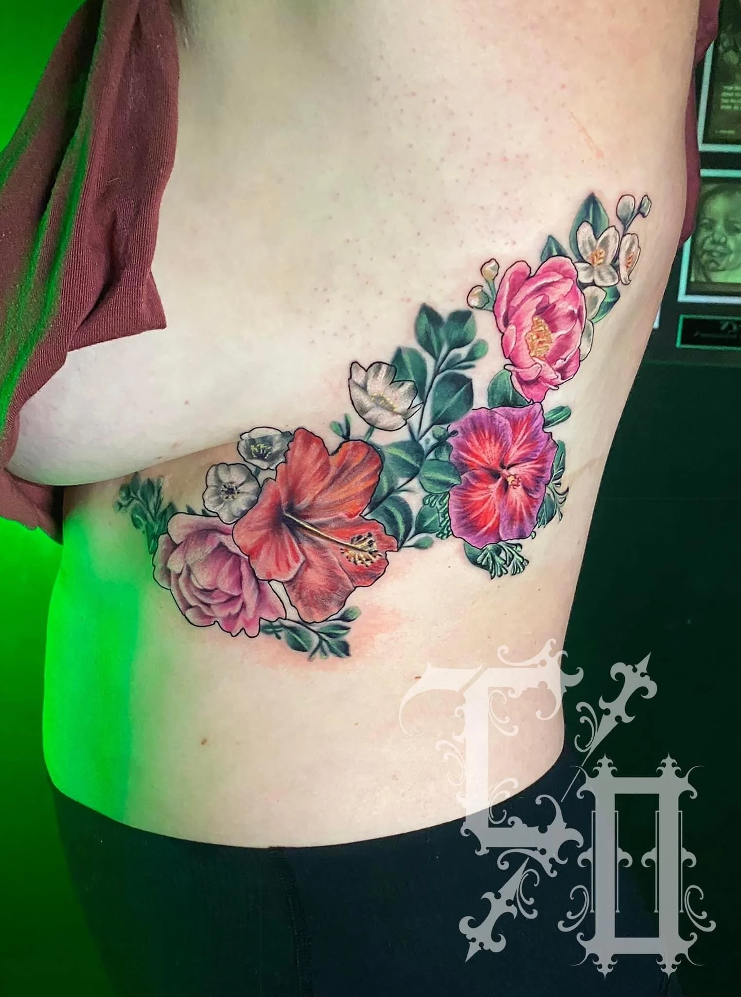 A tattoo of vibrant pink, red, and white hibiscus flowers with green leaves on a person's side torso.