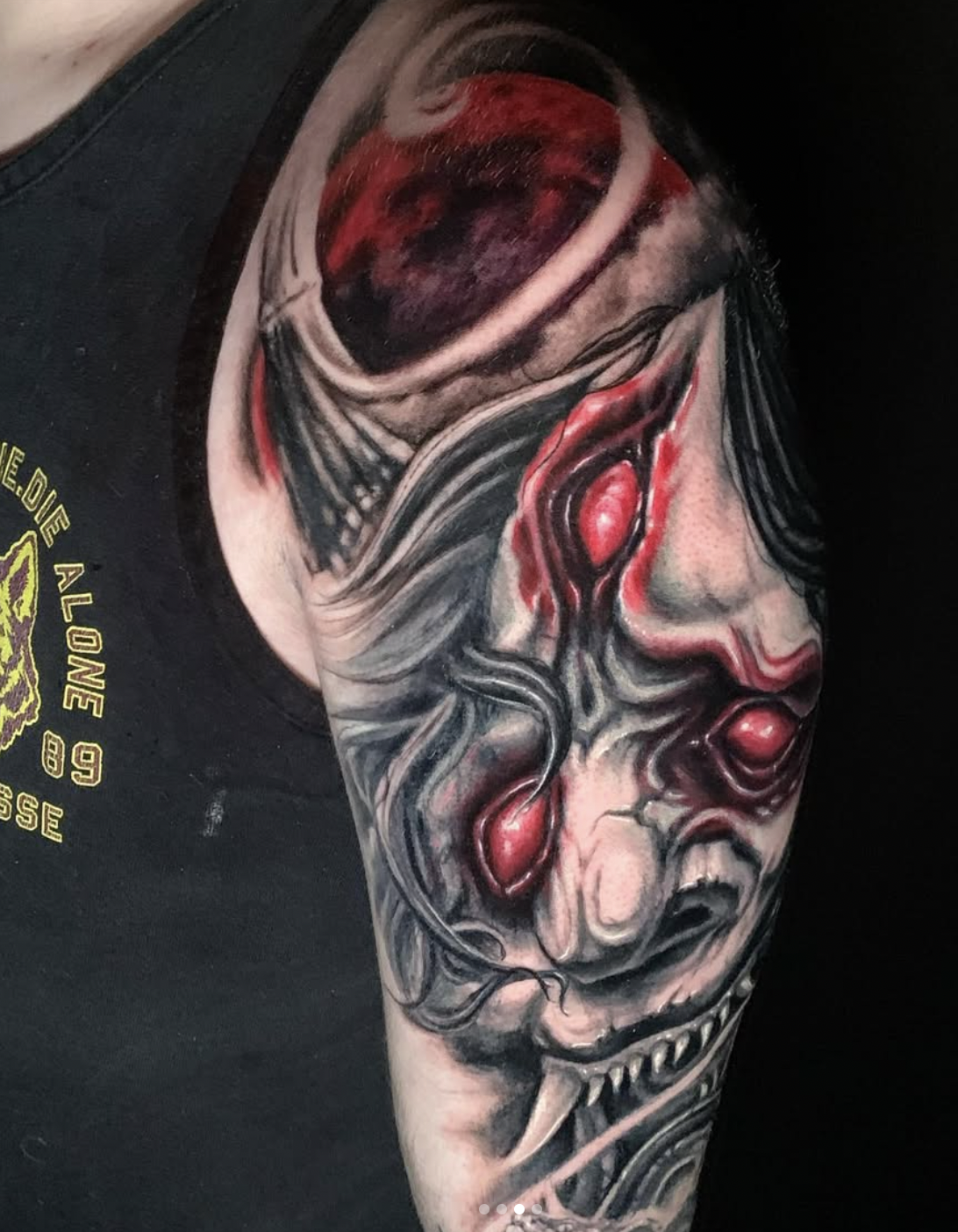 A detailed black, gray, and red tattoo of a screaming clown face with sharp teeth, a red nose, and distorted facial features on a person's upper arm.