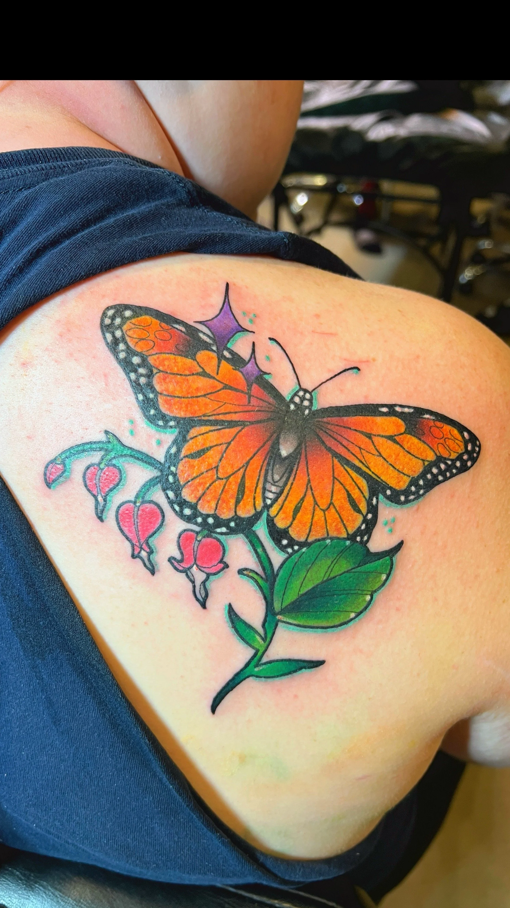 a colorful tattoo with an orange monarch butterfly with red flowers, purple stars, and green leaves behind it on the back of a shoulderblade