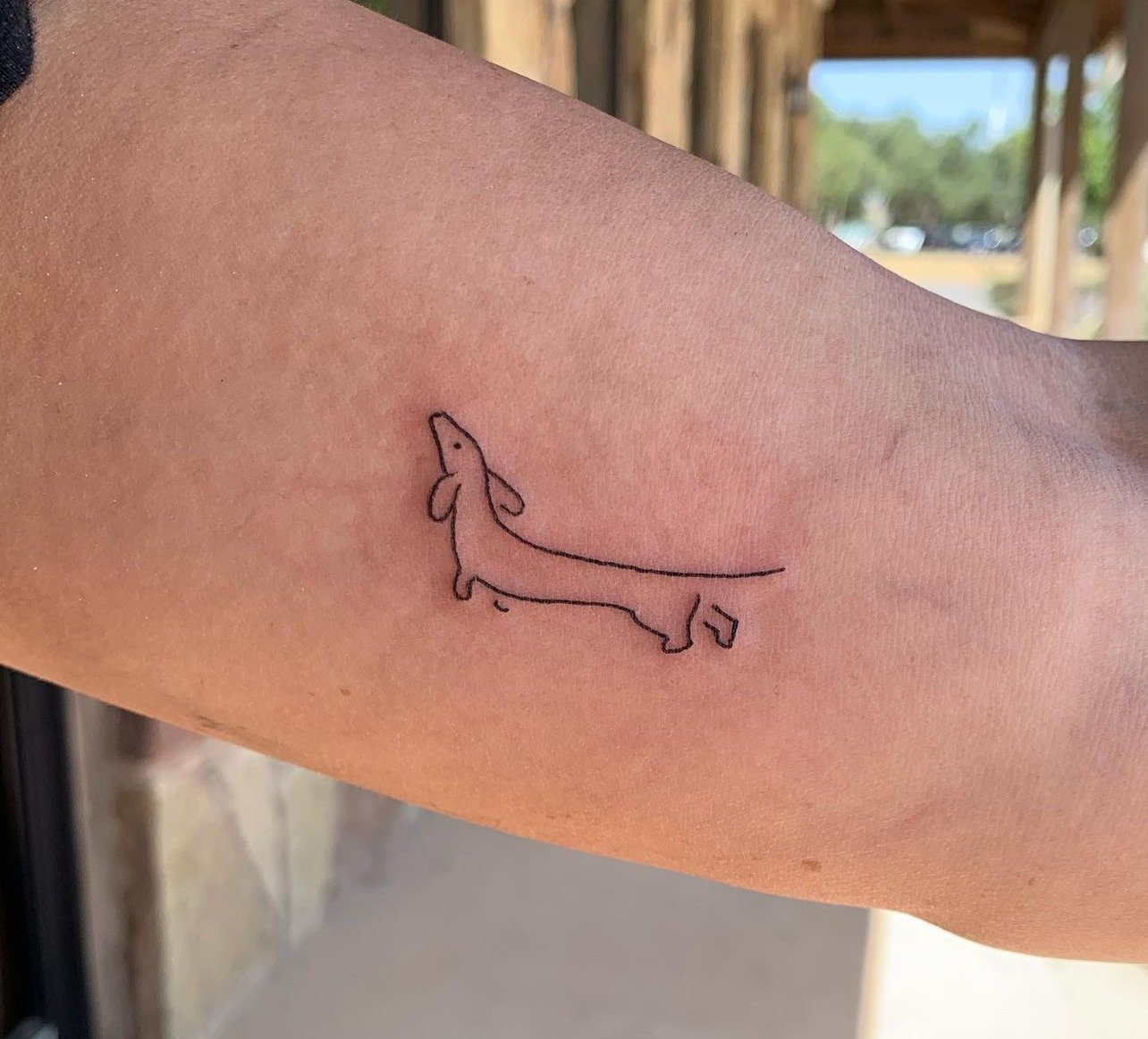 Minimalist tattoo of a dachshund dog outline on a person's arm.