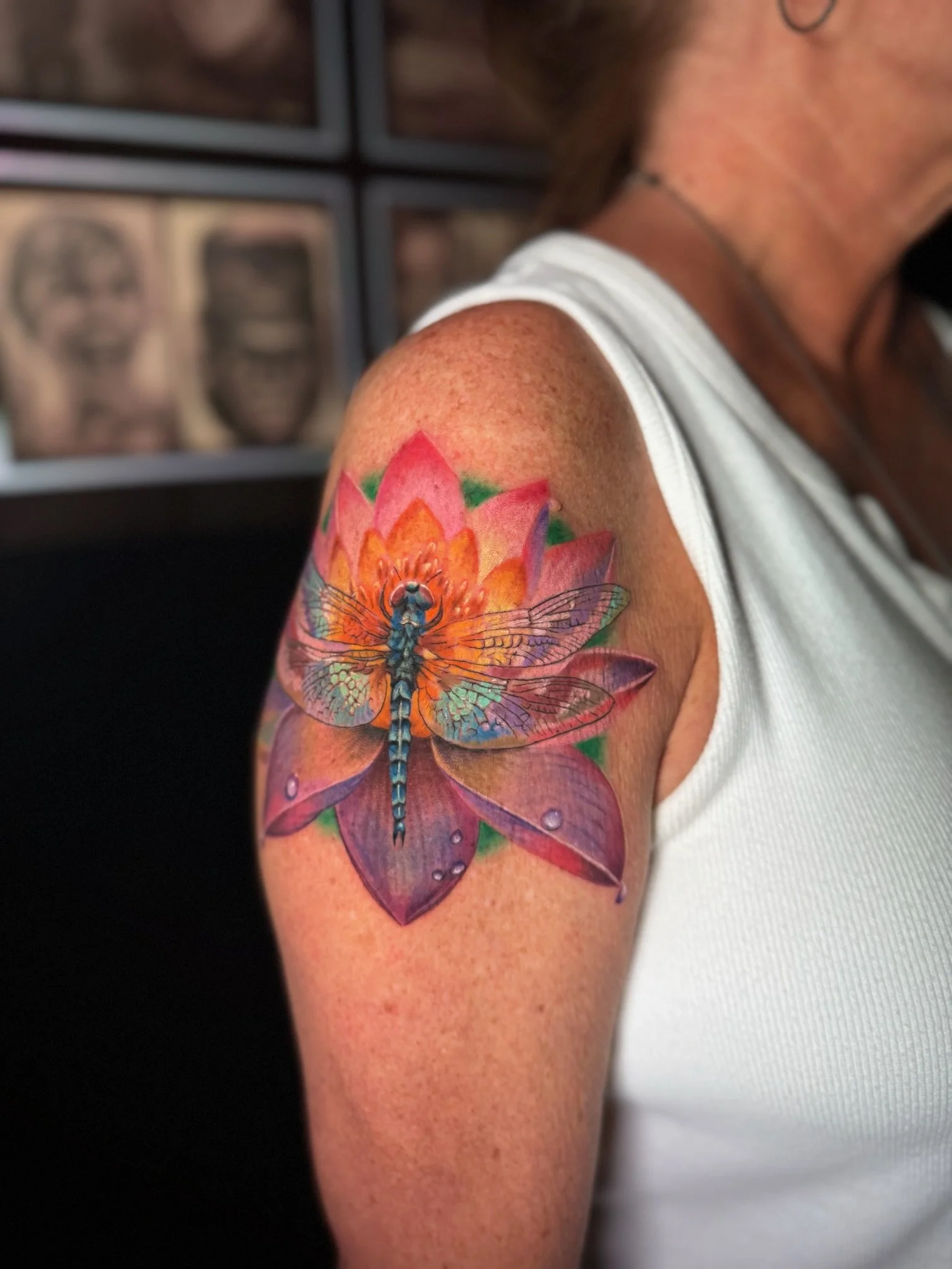 Colorful tattoo of a dragonfly with orange, pink, purple, and blue wings, positioned on a pink lotus flower, on a person's upper arm.